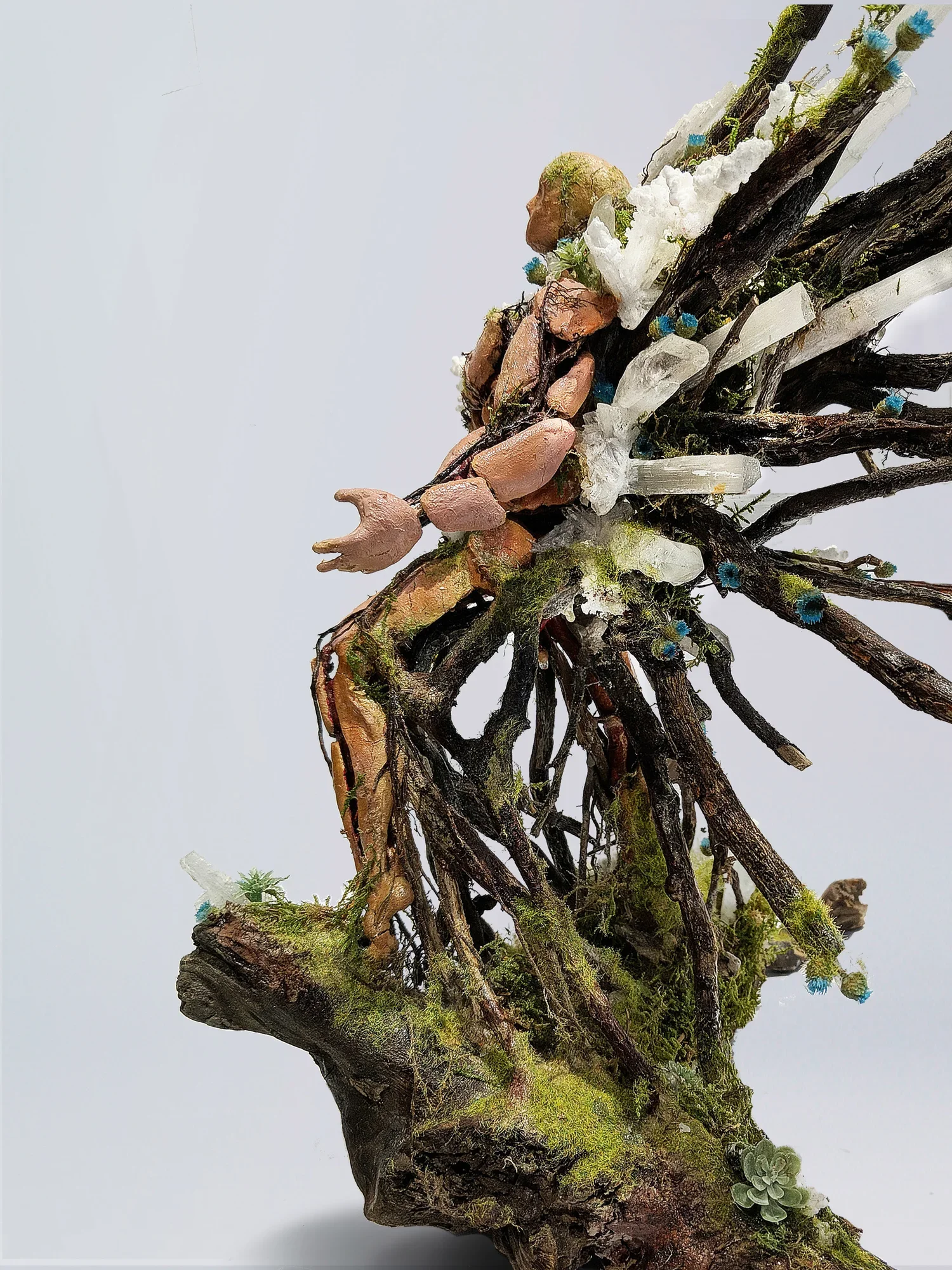 A miniature sculpture of a fairy sitting on a moss-covered branch with small plants and white clouds, against a plain white background.
