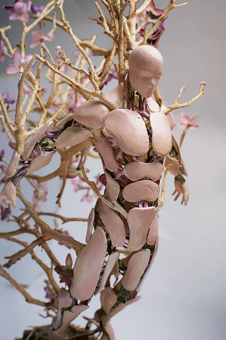 A sculpture of a human figure made from pinkish clay or similar material, integrated with tree branches and pink flowers, creating a hybrid of a person and nature.