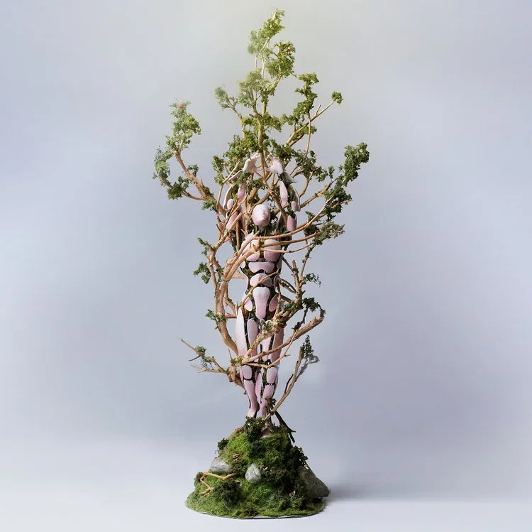 A sculpture of a human figure intertwined with tree branches, with moss at the base and a light background.