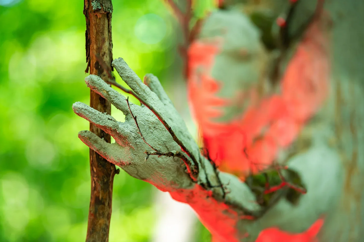Close-up of a textured, pale green hand sculpture with red accents, gripping a brown tree branch against a blurred green background.