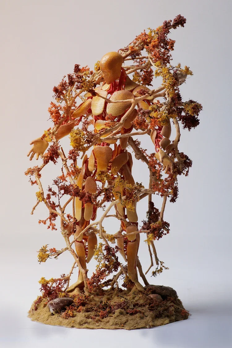 An abstract sculpture of a human figure made with natural materials, including small branches, leaves, and organic objects, resembling a person wrapped in a tree or foliage.