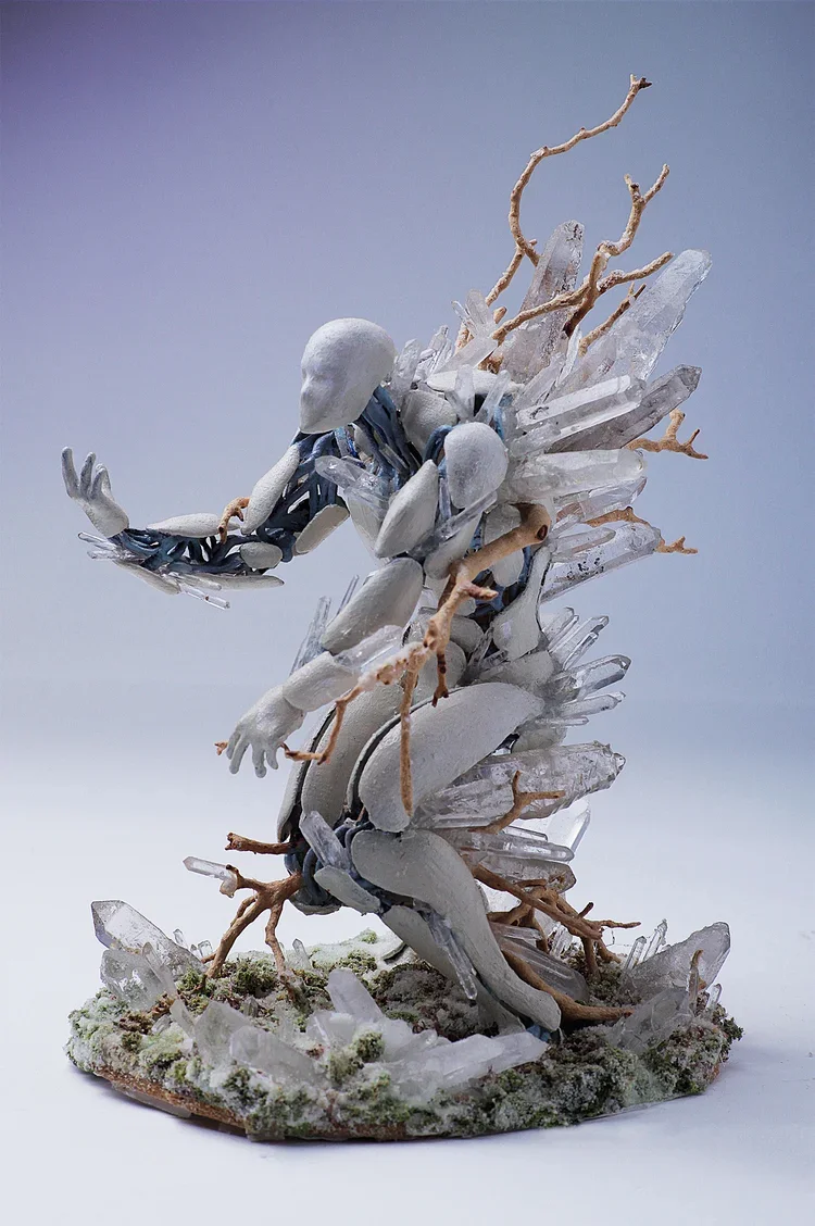 A miniature sculpture of a human figure surrounded by white crystals and brown branches, set on a base with moss and smaller crystals.