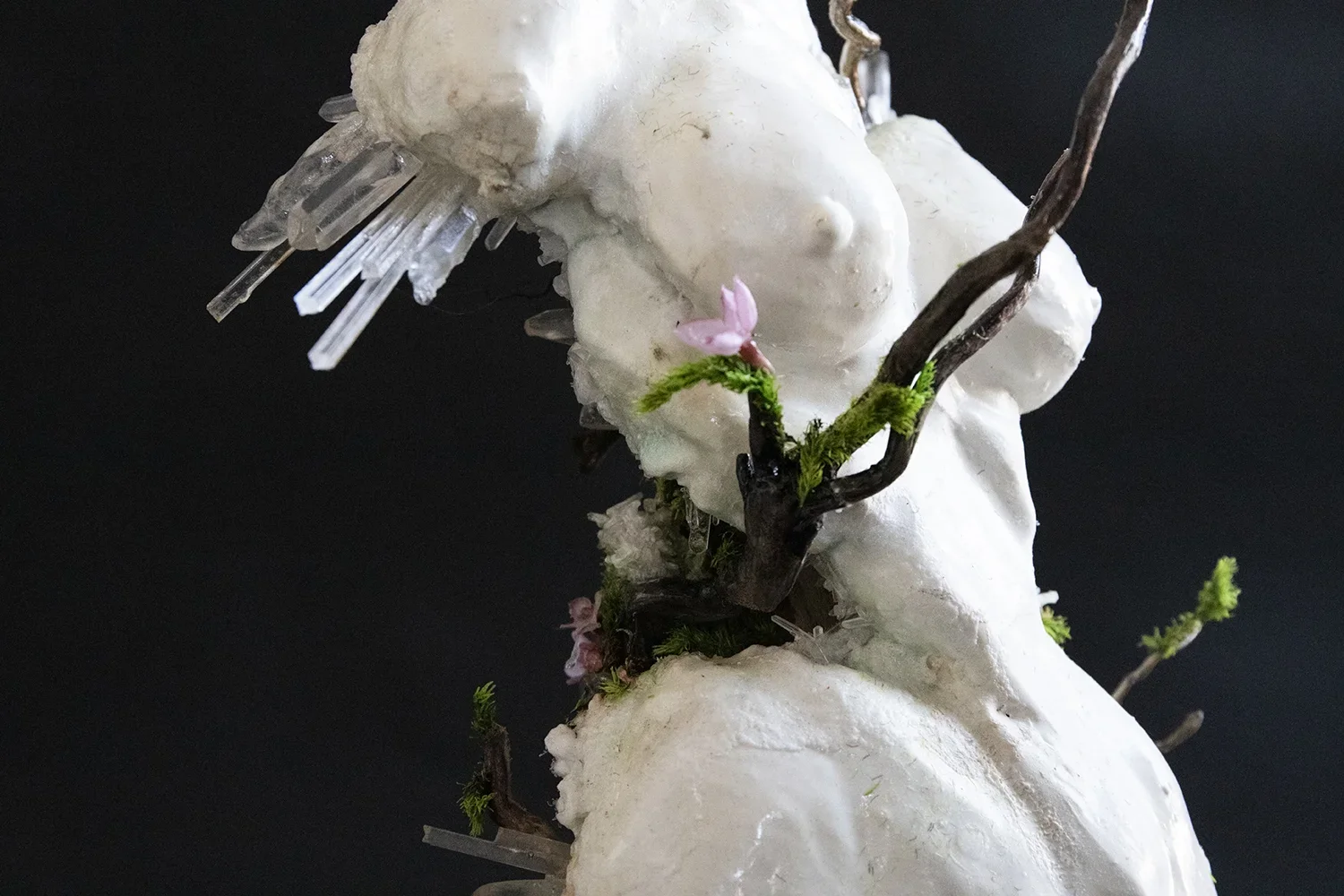 Sculpture of a reclining woman with closed eyes, made of white material, adorned with small pink flowers, green moss, and brown twig-like structures, set against a black background.