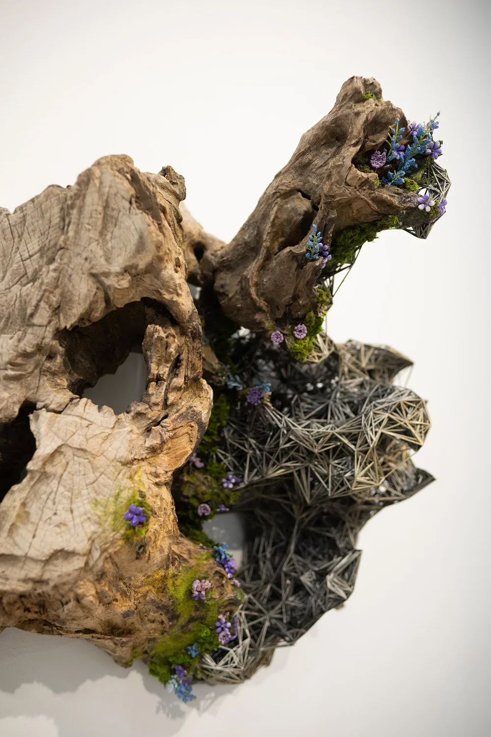 Mixed media art piece combining natural wood and geometric metal structures with small purple, blue, and green flowers and moss.