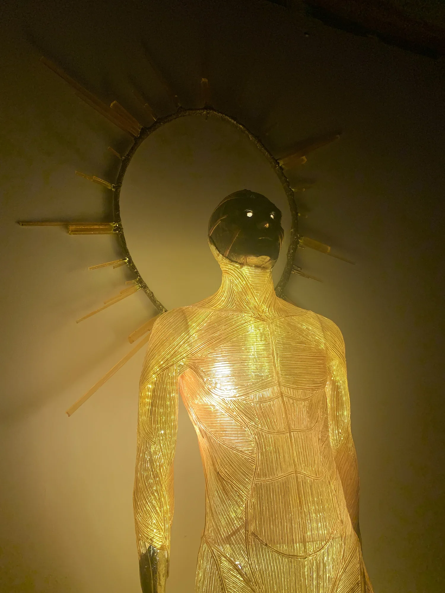 Golden statue of a woman with a lid-like structure surrounding her head, reminiscent of a halo, with internal lighting highlighting her form.