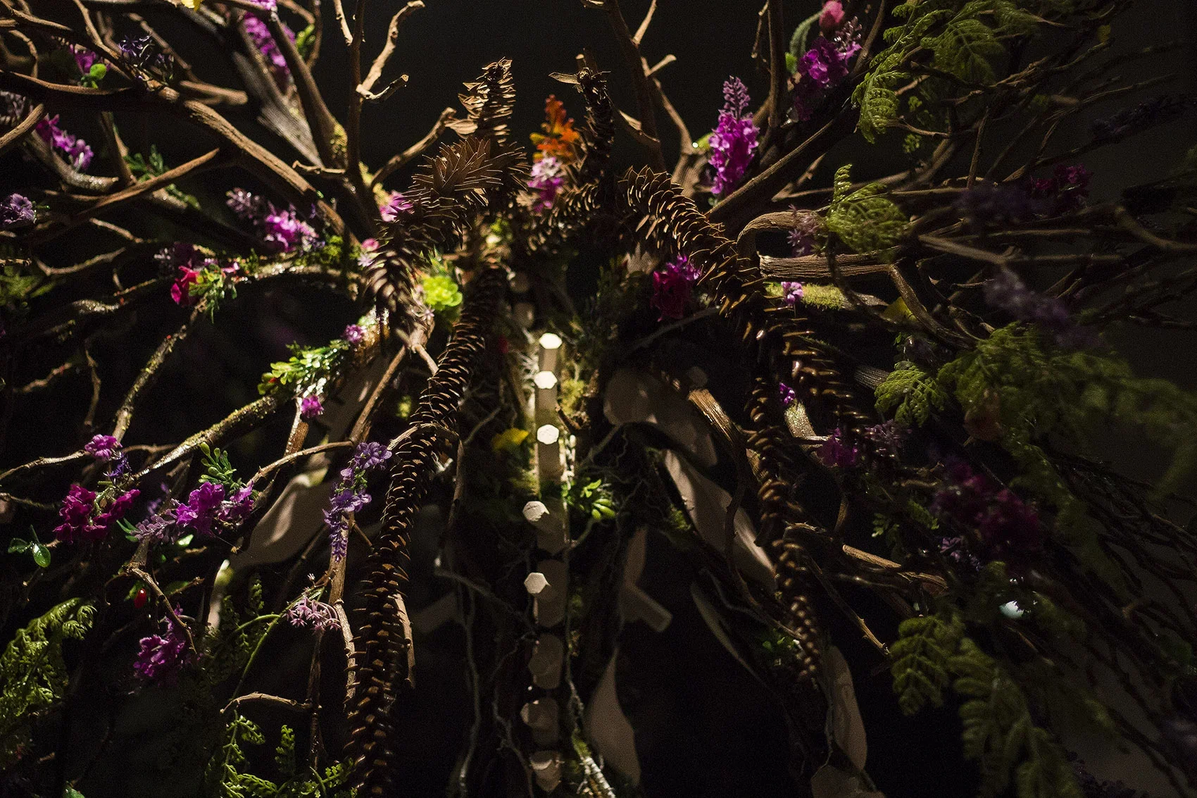 Darkly lit arrangement of intertwined branches, green moss, and purple flowers, with small white lights illuminating the scene.