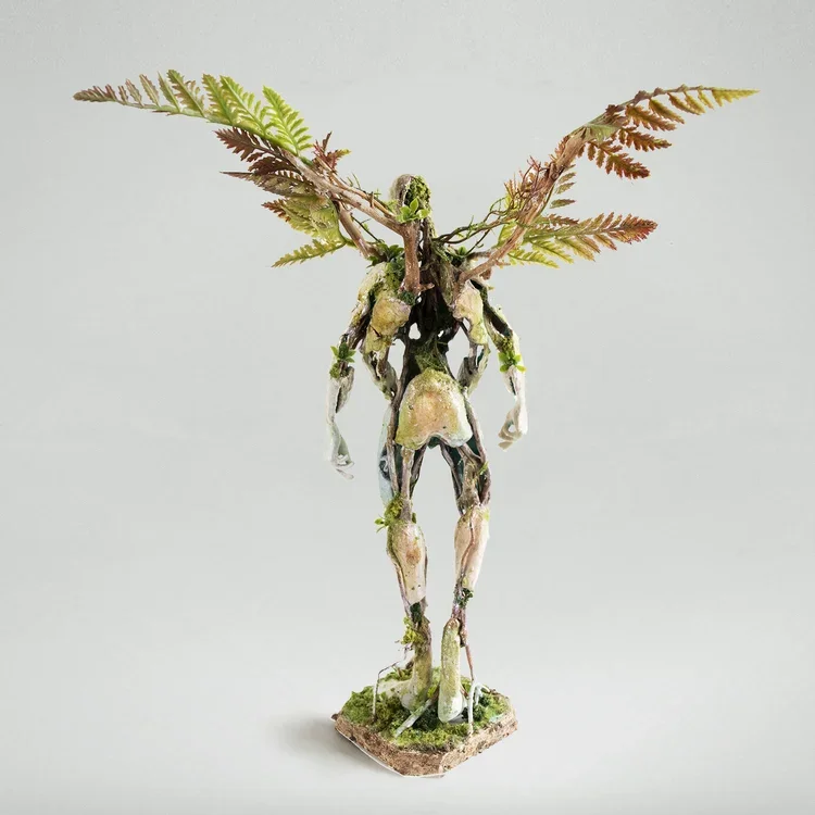 A small sculpture of a humanoid figure made from natural materials, with fern leaves for wings, standing on a base of dirt or bark.