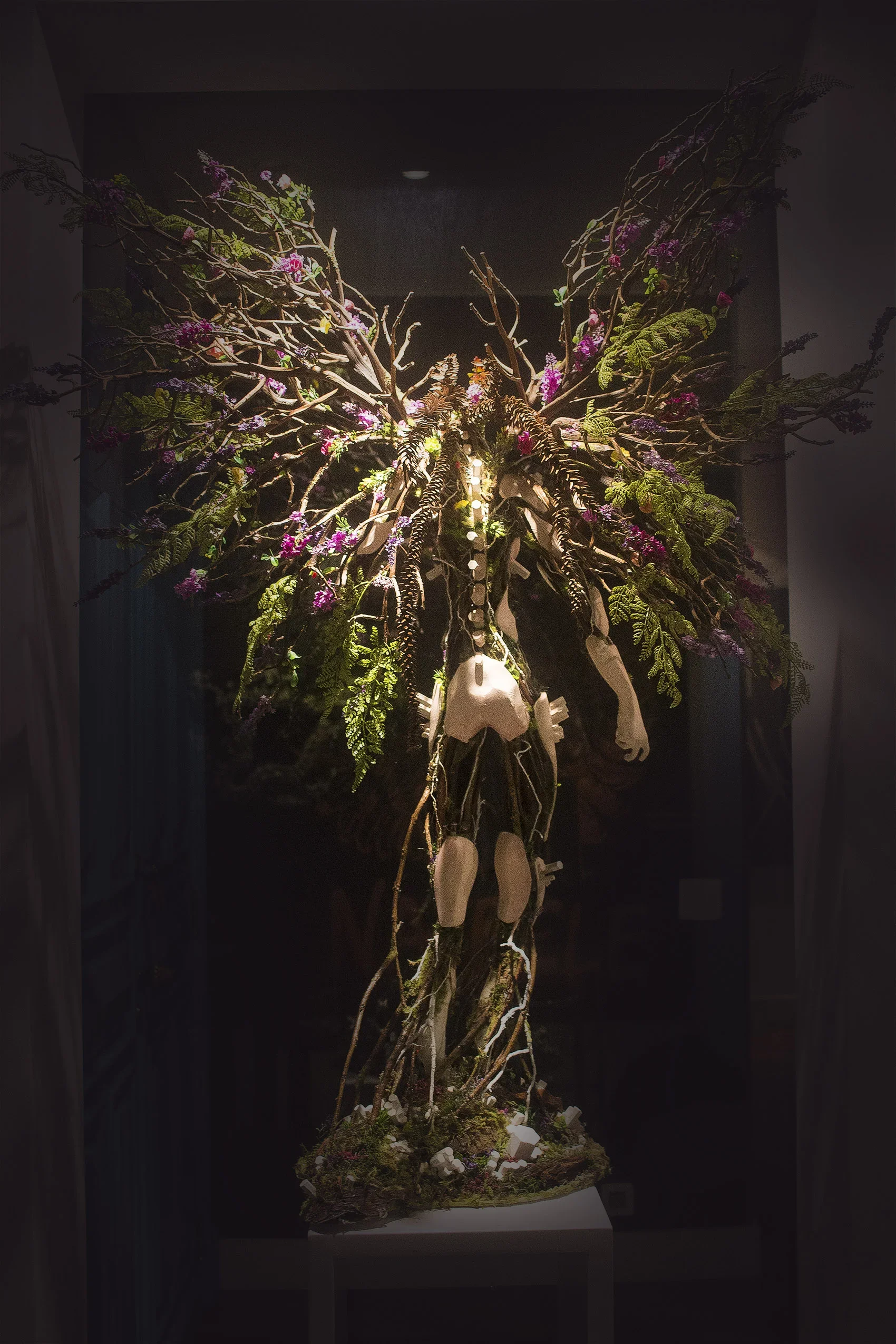 A sculpture of a human skeleton intertwined with a tree, flowers, and branches, displayed indoors with dramatic lighting.