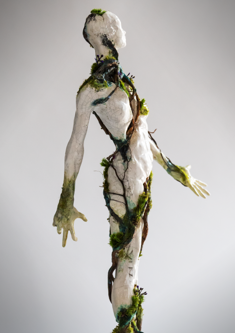 A humanoid sculpture with a cracked white surface, covered in moss and vines, standing against a plain, neutral background.