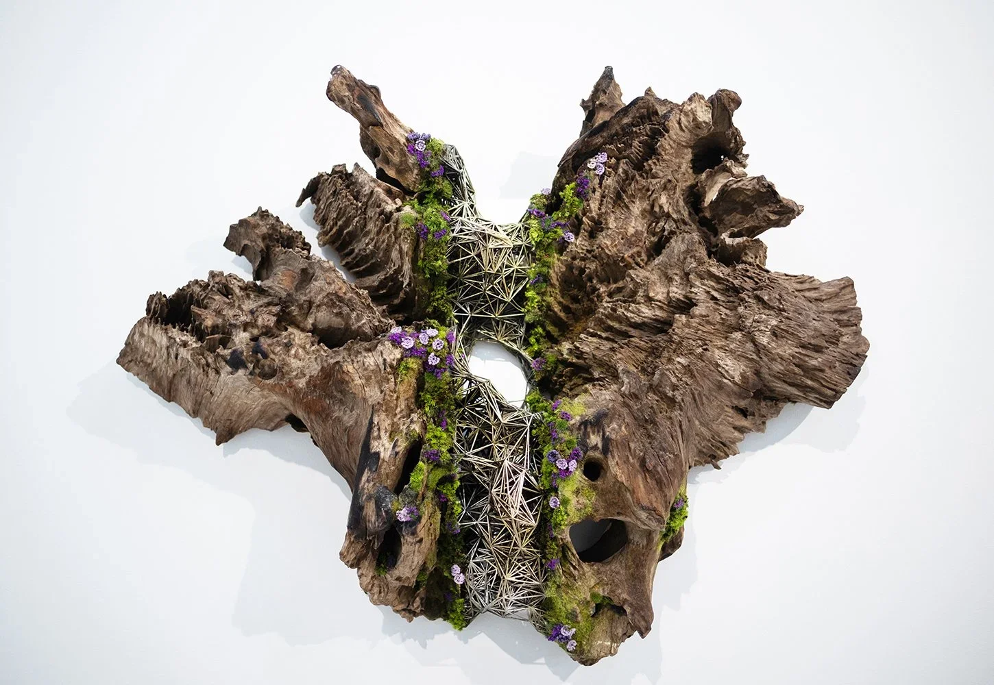 Mixed media art piece featuring a large, textured wooden sculpture with moss and purple flowers, accented by geometric metal framework.