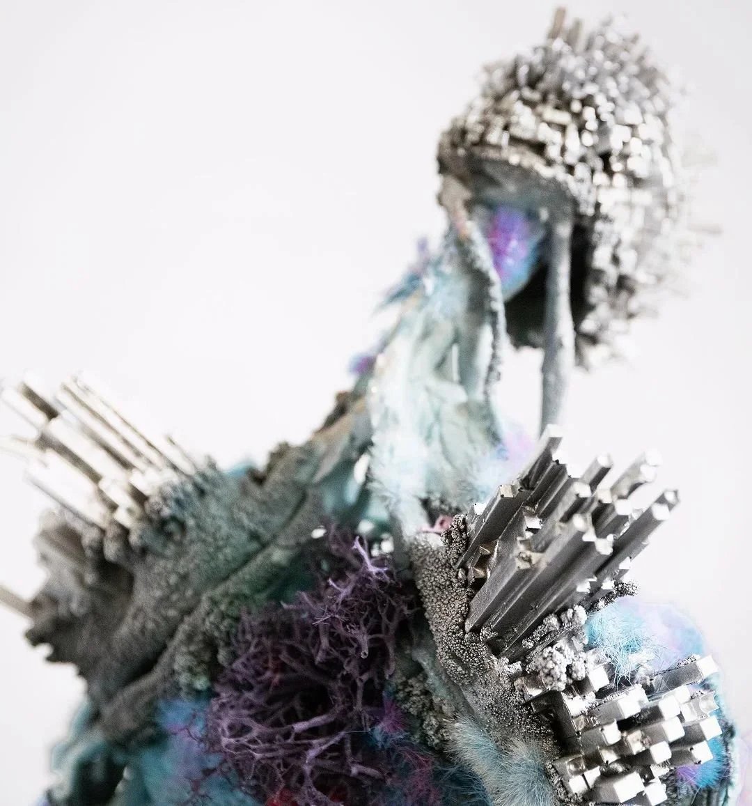 Abstract sculpture featuring metallic and colorful textured materials, including silver spikes, purple and blue fibers, and a humanoid face with a headdress.