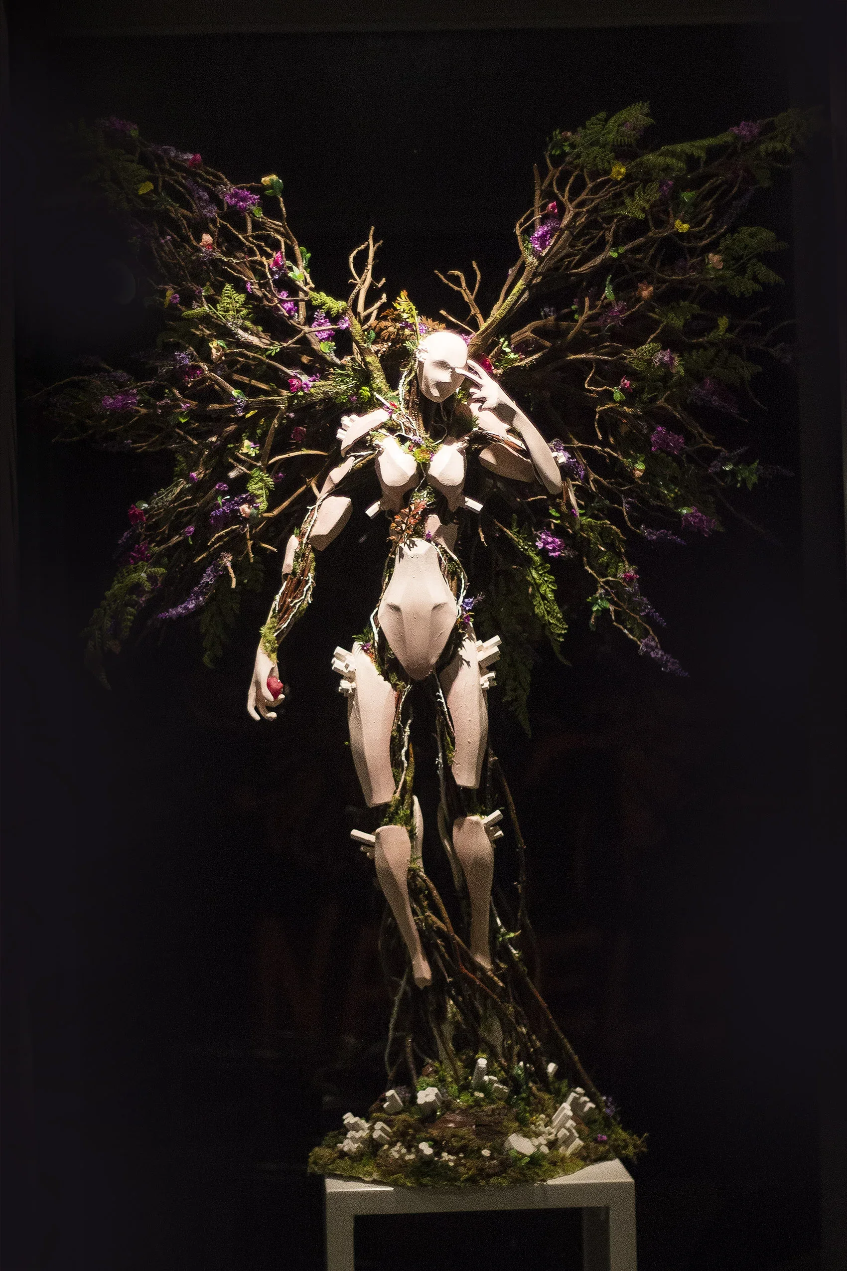 A sculpture of a human figure with exaggerated features, decorated with intertwining branches and colorful flowers, displayed on a pedestal against a dark background.