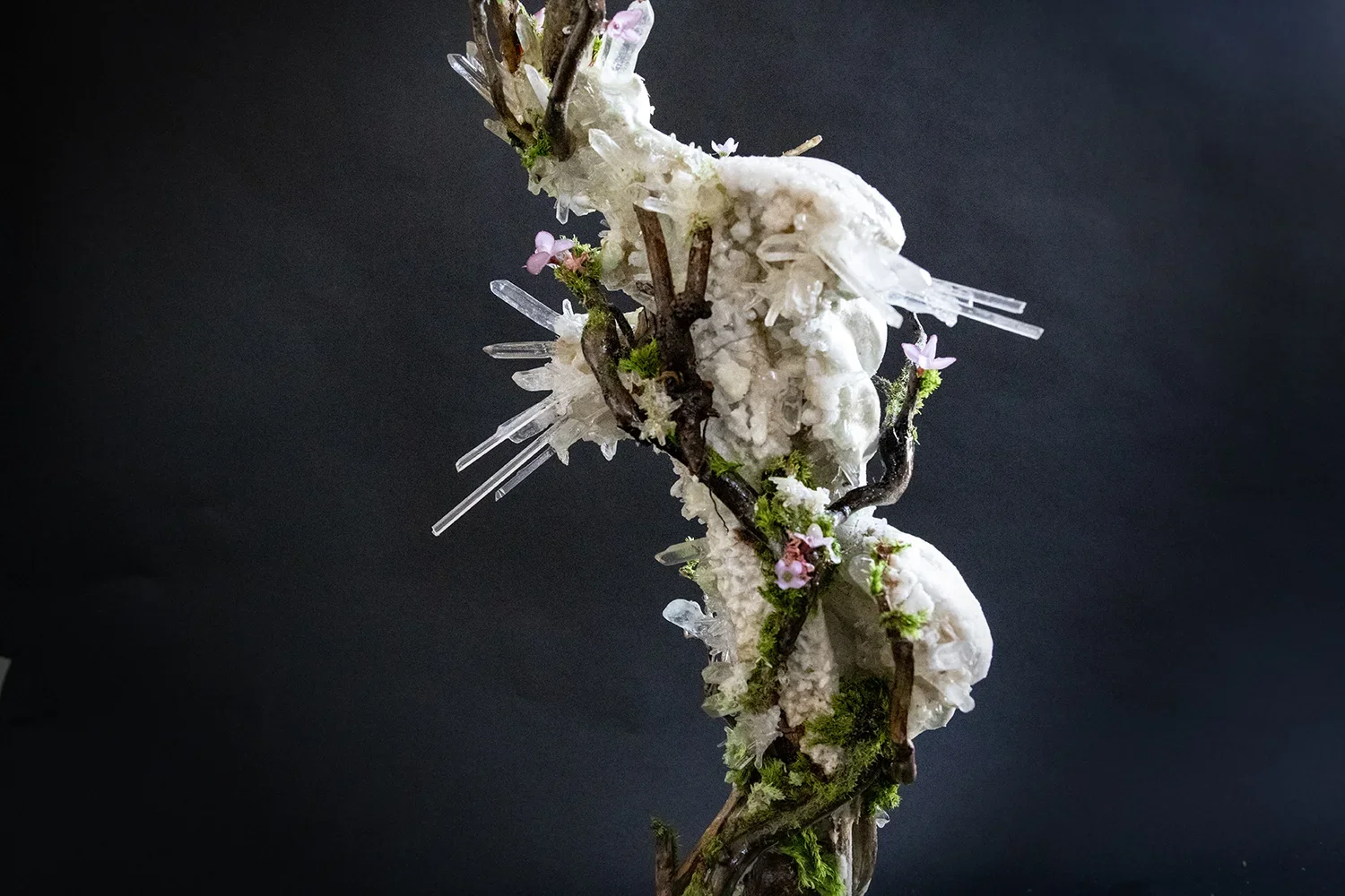 An abstract art sculpture featuring twisted branches, white textured materials, pink flowers, green moss, and clear glass-like elements against a dark background.
