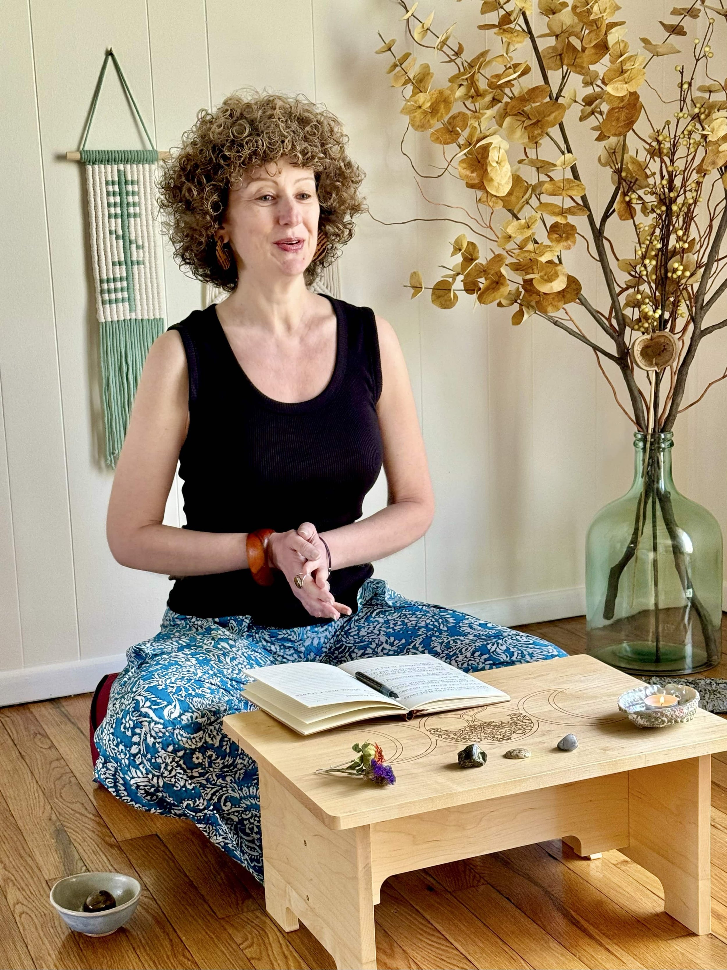 A woman with curly hair sitting on the floor behind a small wooden table, with an open book and writing instrument, in a room with dried flowers, a large glass vase with branches and leaves, and a wall hanging.
