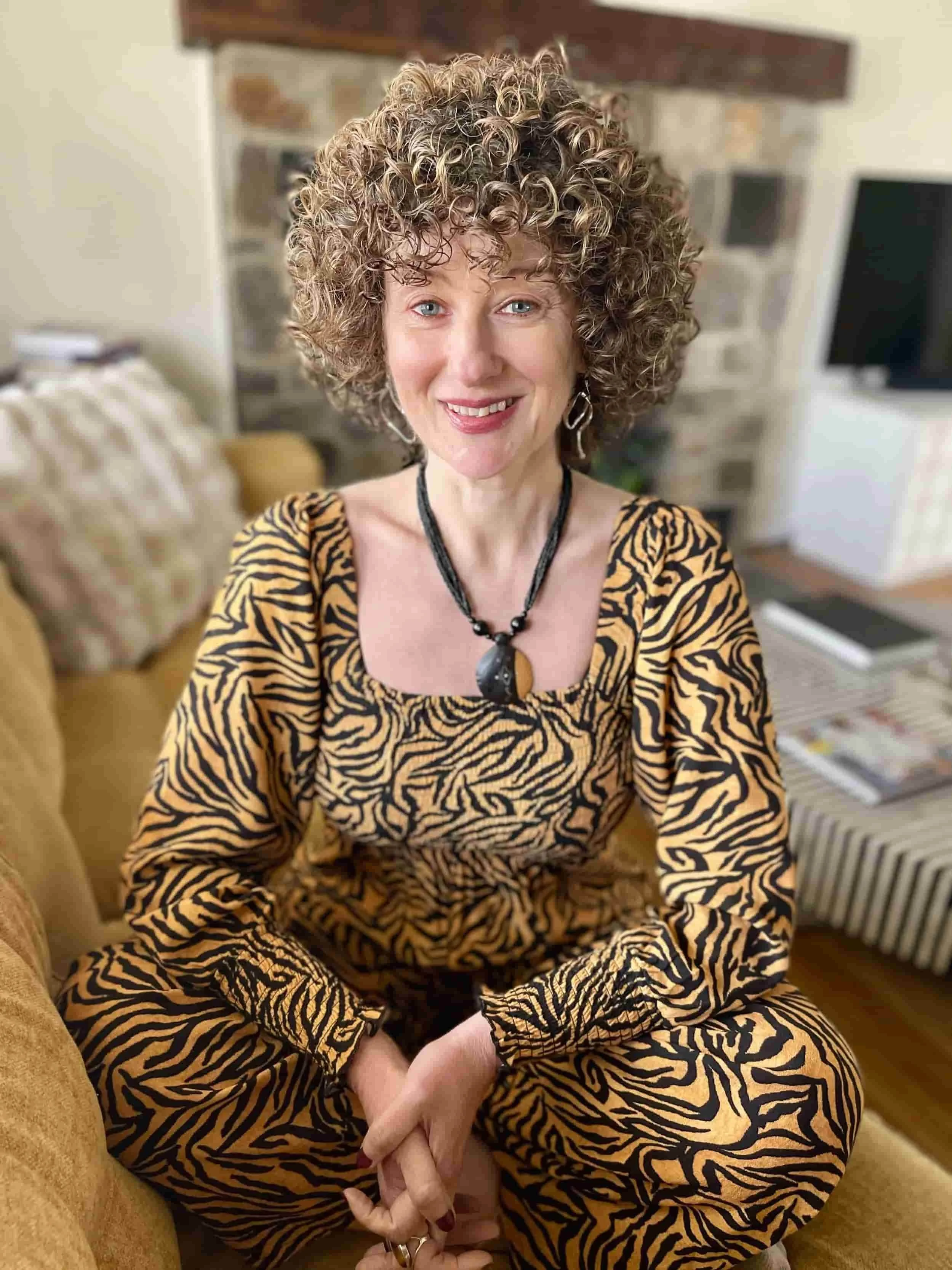 Smiling woman with curly hair in a tiger-striped outfit sits cross-legged on a couch in a cozy living room.