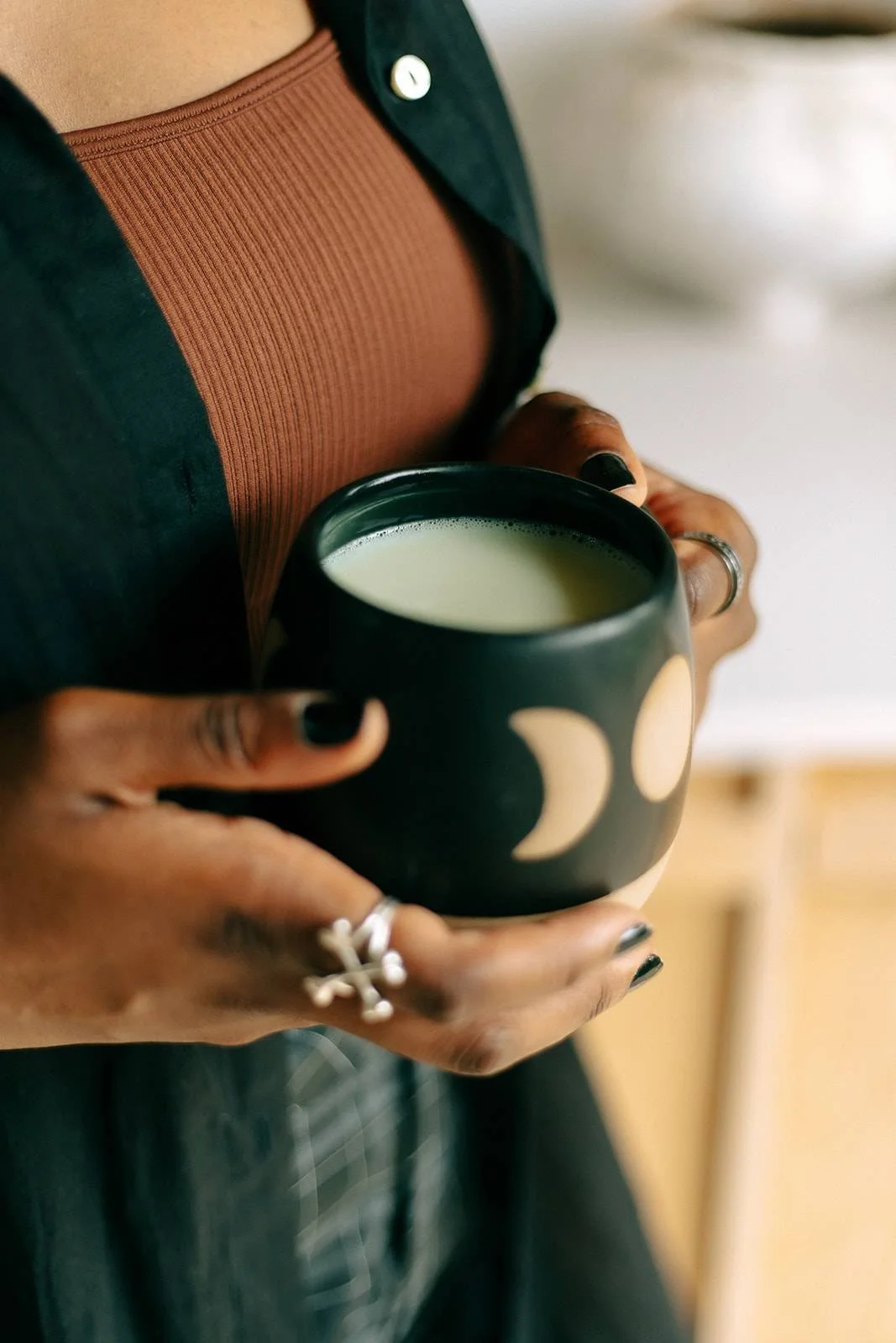 Dominique holding a black mug with moon and crescent moon designs, filled with matcha tea. She's wearing rings, a dark cardigan, and a brown ribbed top.
