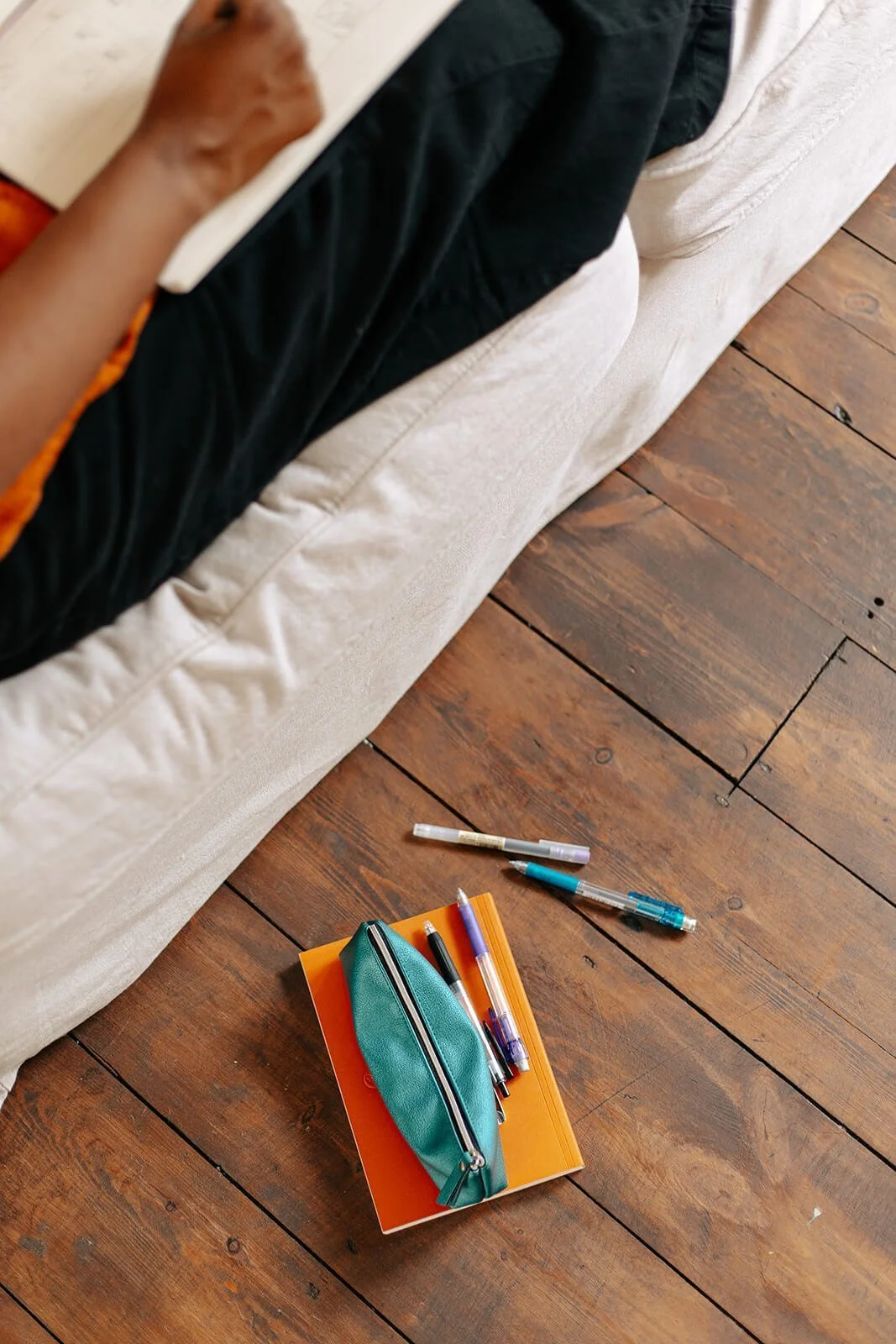 A beige couch with various pens and a teal pencil case on the wooden floor beside it.