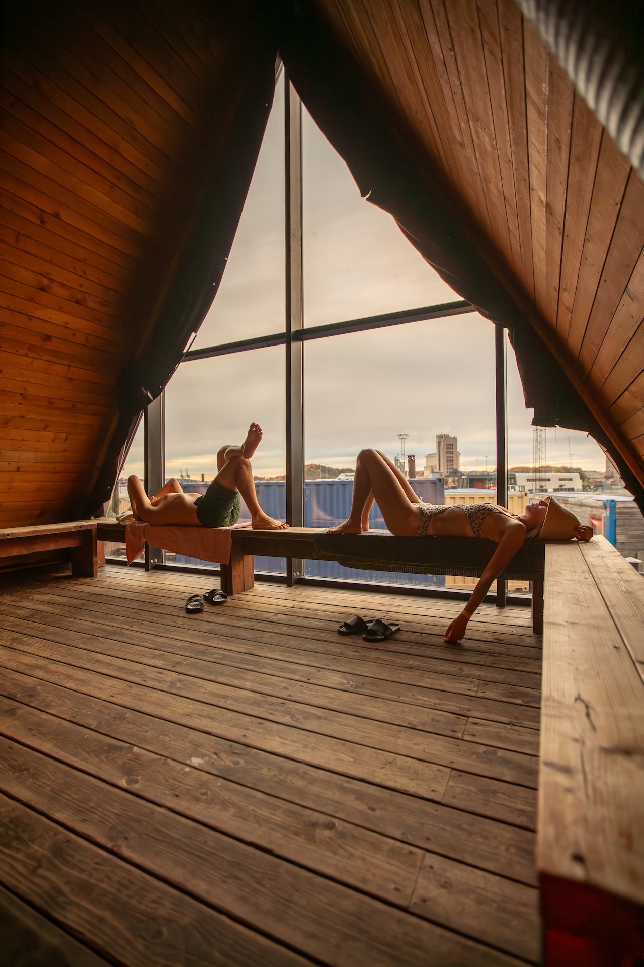 Two people lounge on a bench inside a wooden A-frame cabin with large floor-to-ceiling windows, overlooking a cityscape.