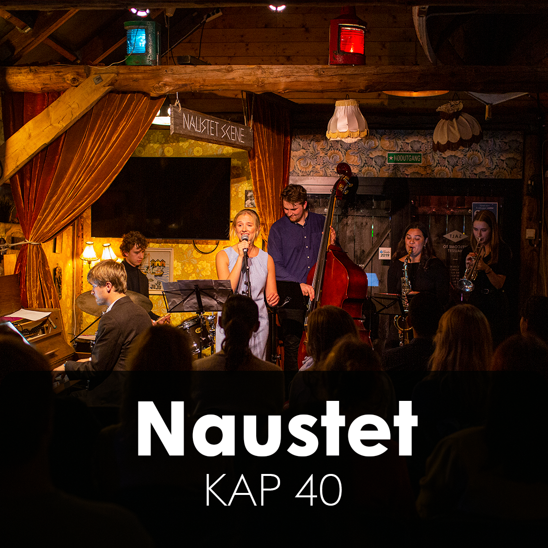 A group of women singing on stage in a cozy, dimly lit bar or cafe, with a decorated interior and audience watching.