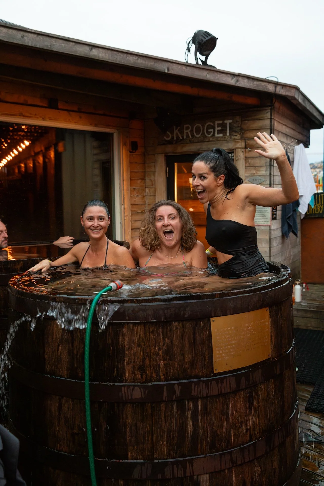 Three women smiling and laughing in a hot tub outside a wooden building with the sign 'SKROGET' visible.