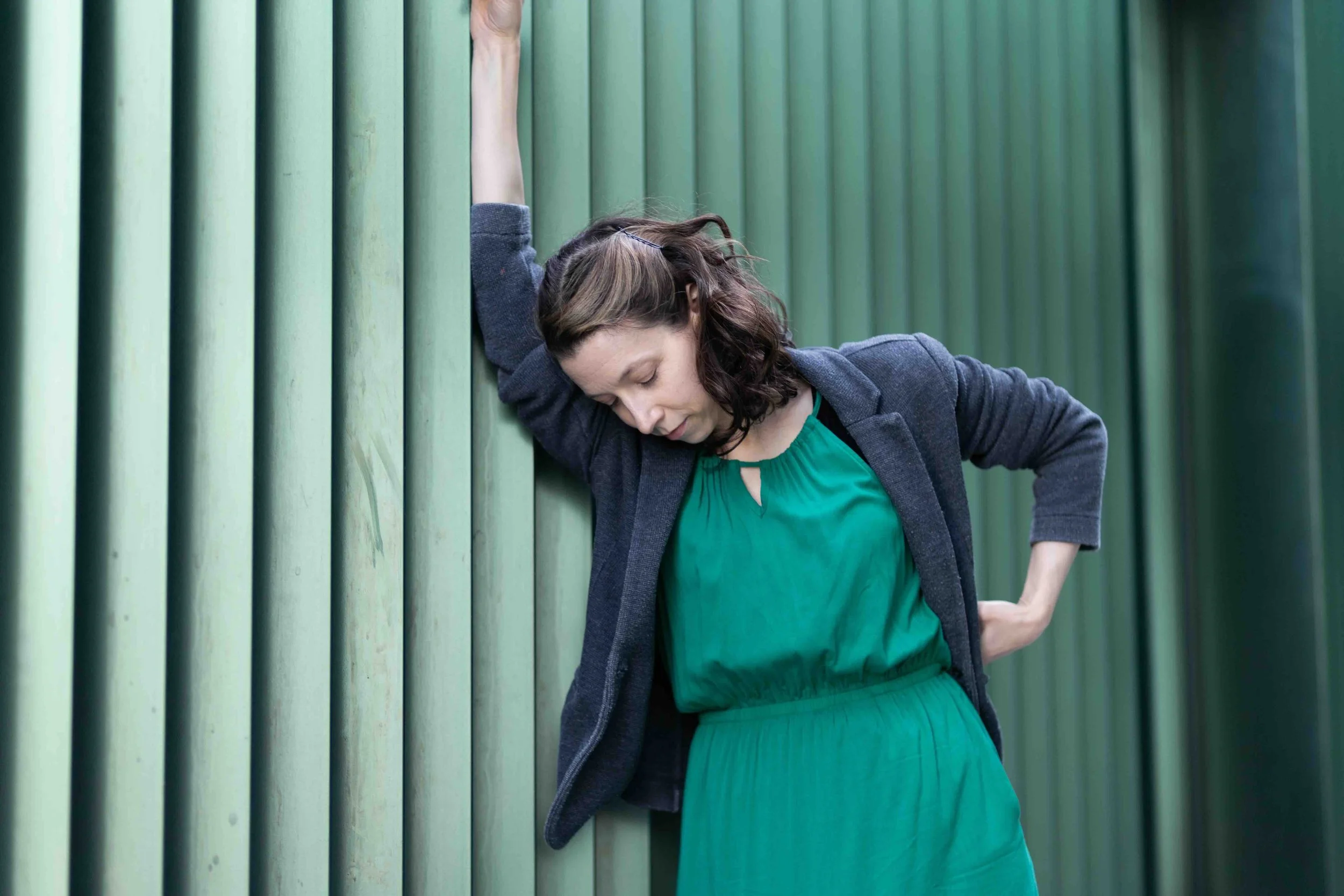 Rowen the actor and singer leans against a green corrugated wall in a long green dress and grey jacket, in an 'I'm a little teapot' pose.