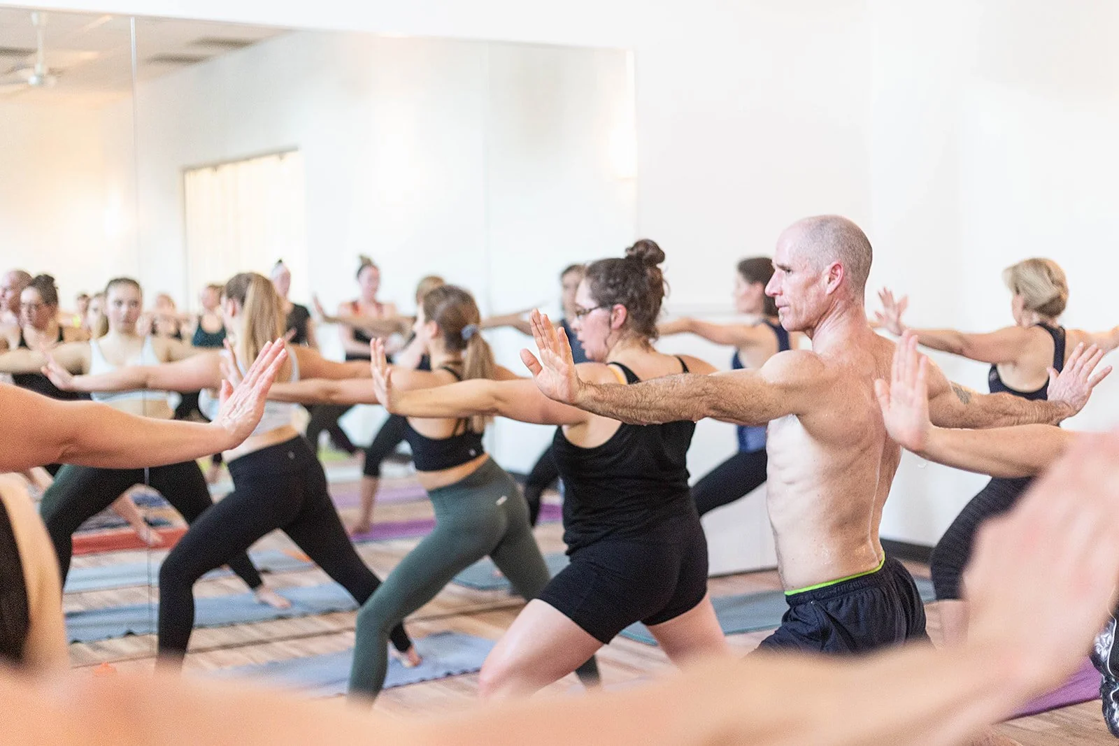 A group of men and womean stand in warrior 1 in a well lit yoga studio