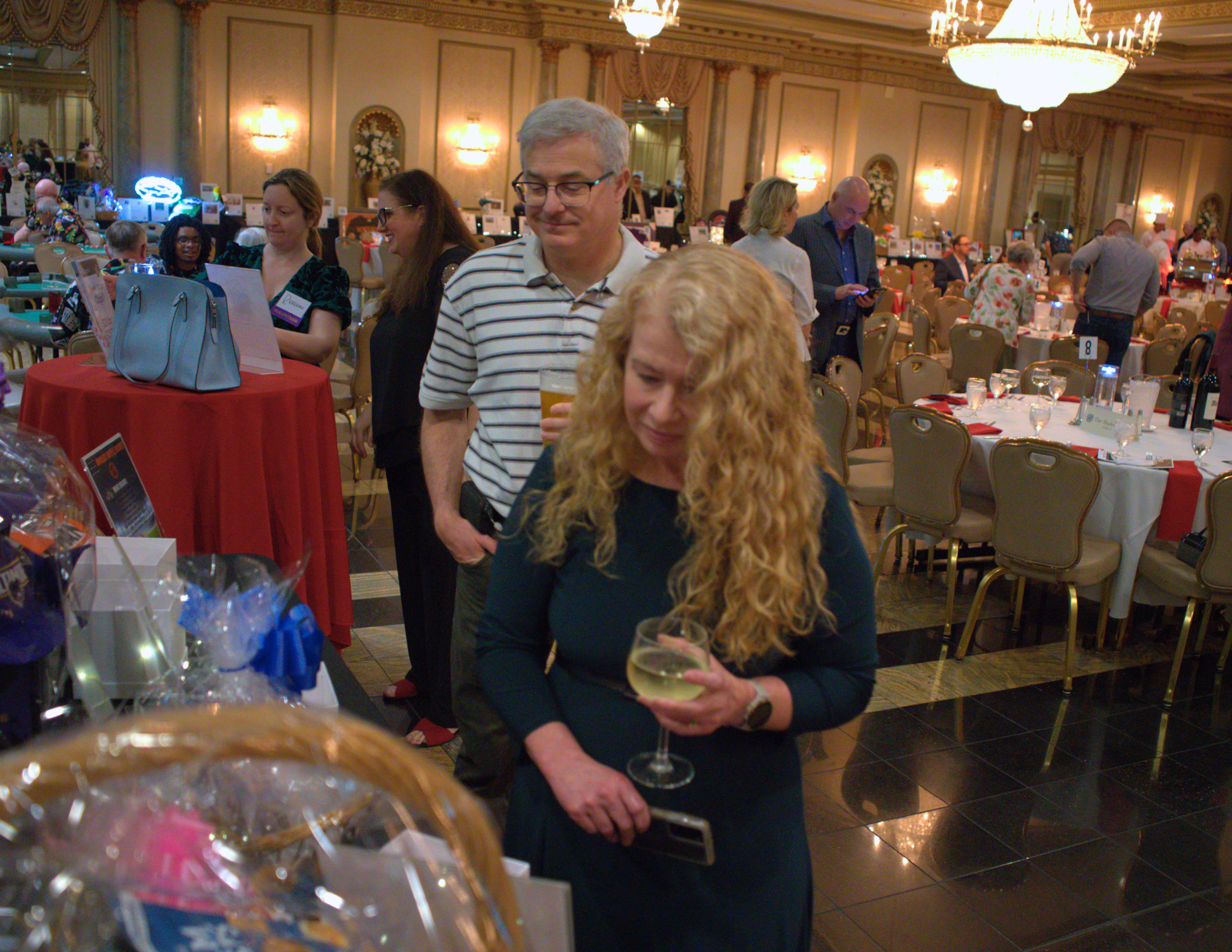 a blonde woman and gray haired man looking at auction items while holding drinks.