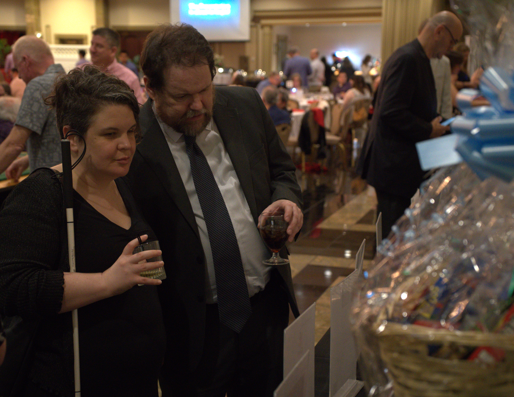 a man with a beard and a blind woman with a cane standing in front of silent auction items.