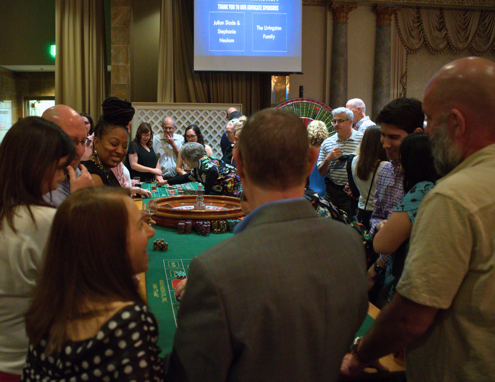 a very large group of all ages people standing at a roulette table and laughing.