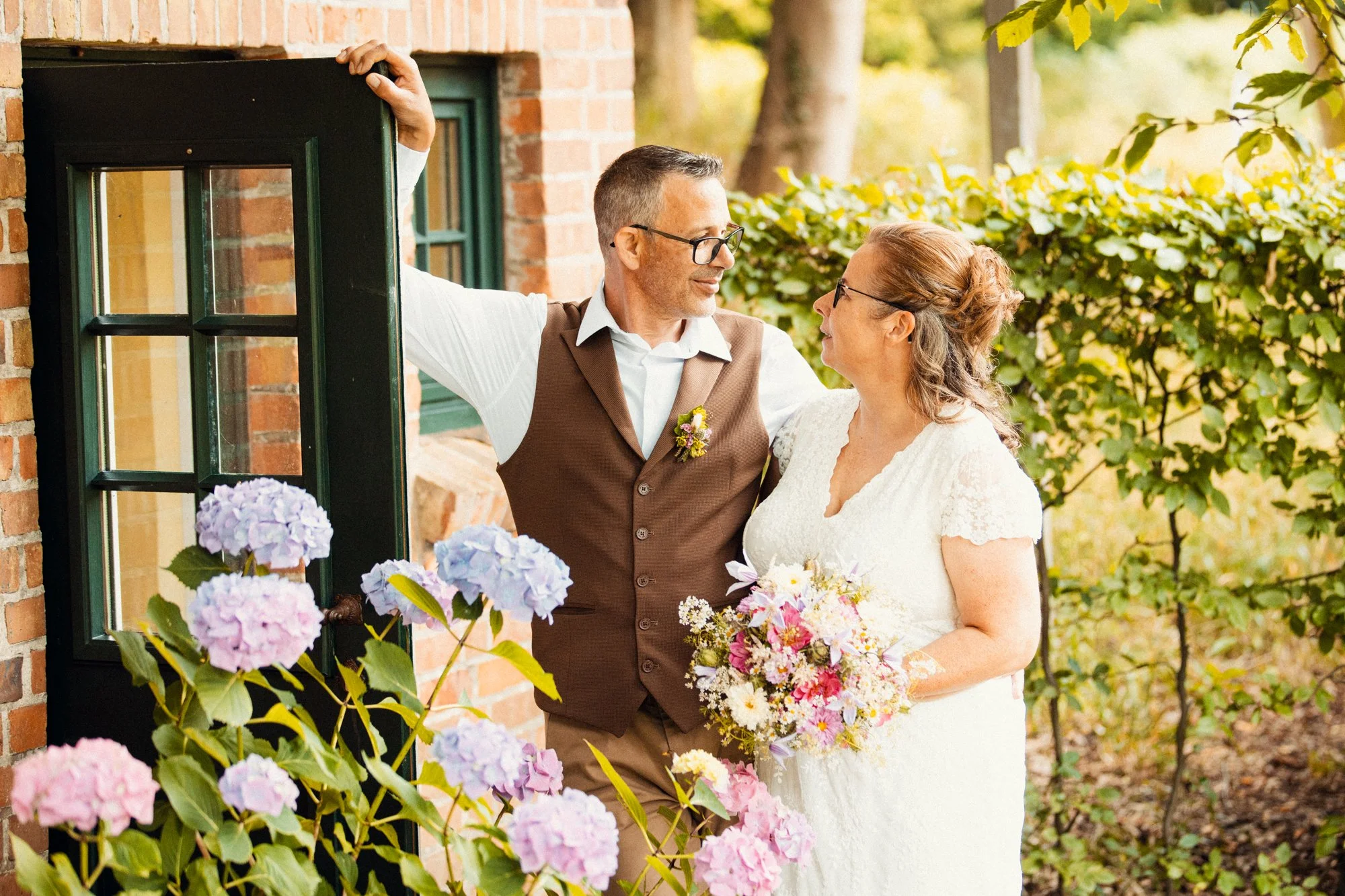 A couple dressed in wedding attire standing outside a brick house, surrounded by flowers and greenery, looking at each other affectionately.