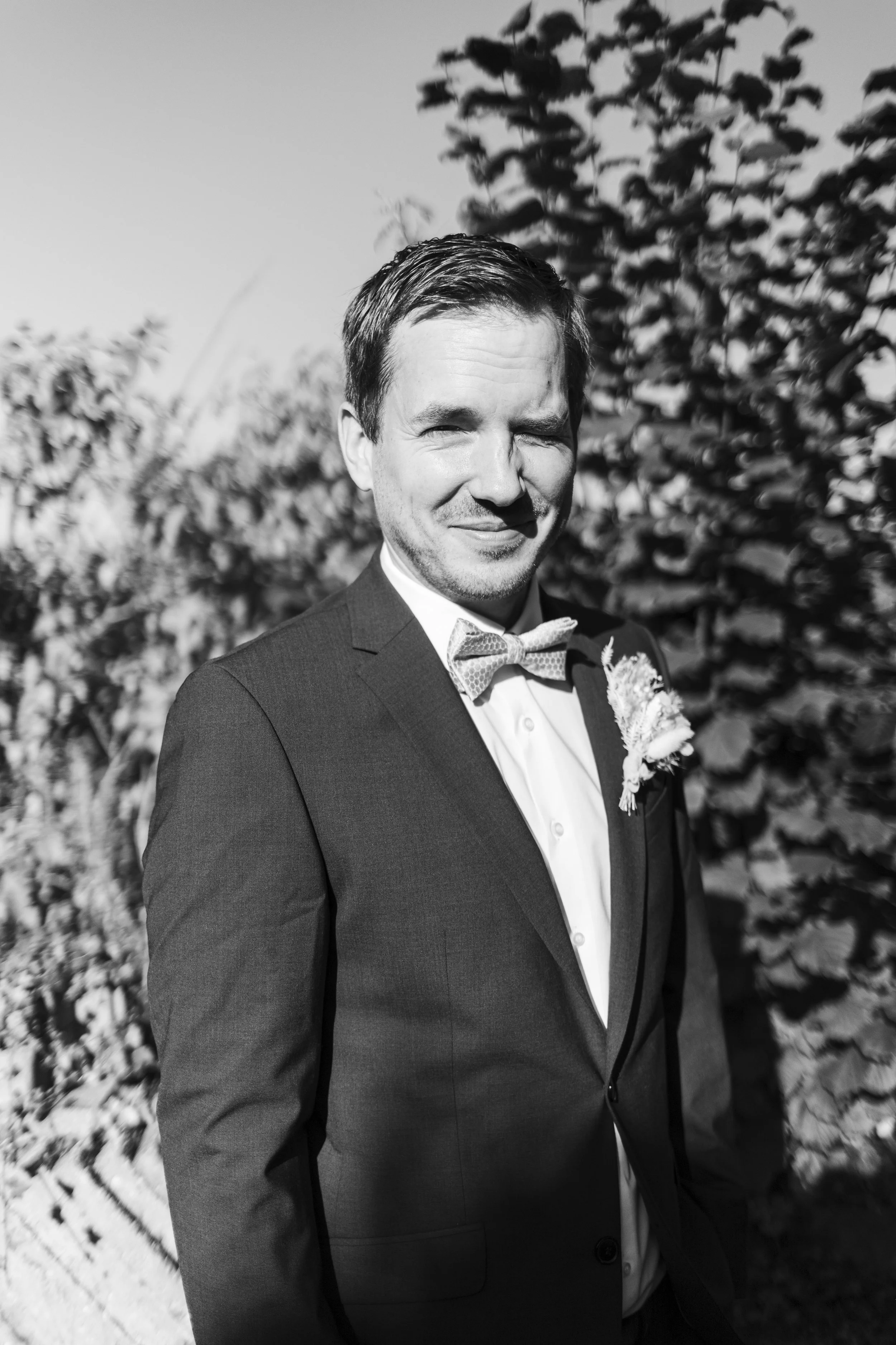 A black and white photo of a man in a tuxedo with a bow tie and boutonniere, smiling with eyes partially closed, standing outdoors with foliage in the background.