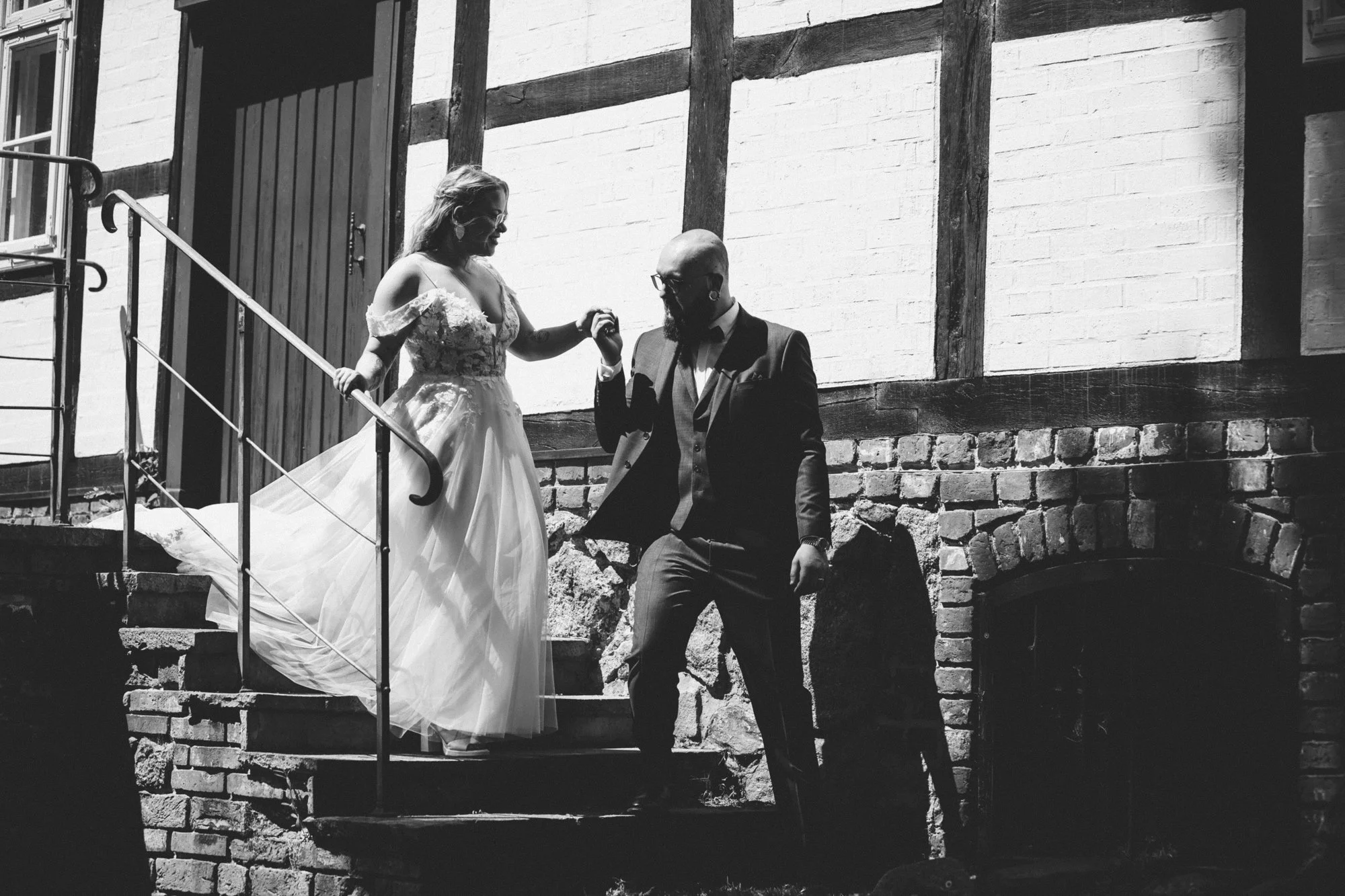 A black and white photo of a bride and groom on a stone and brick staircase outside. The bride, wearing a wedding dress, is holding the groom's hand and looking down at him. The groom, in a suit and tie, is kneeling and looking at the bride."