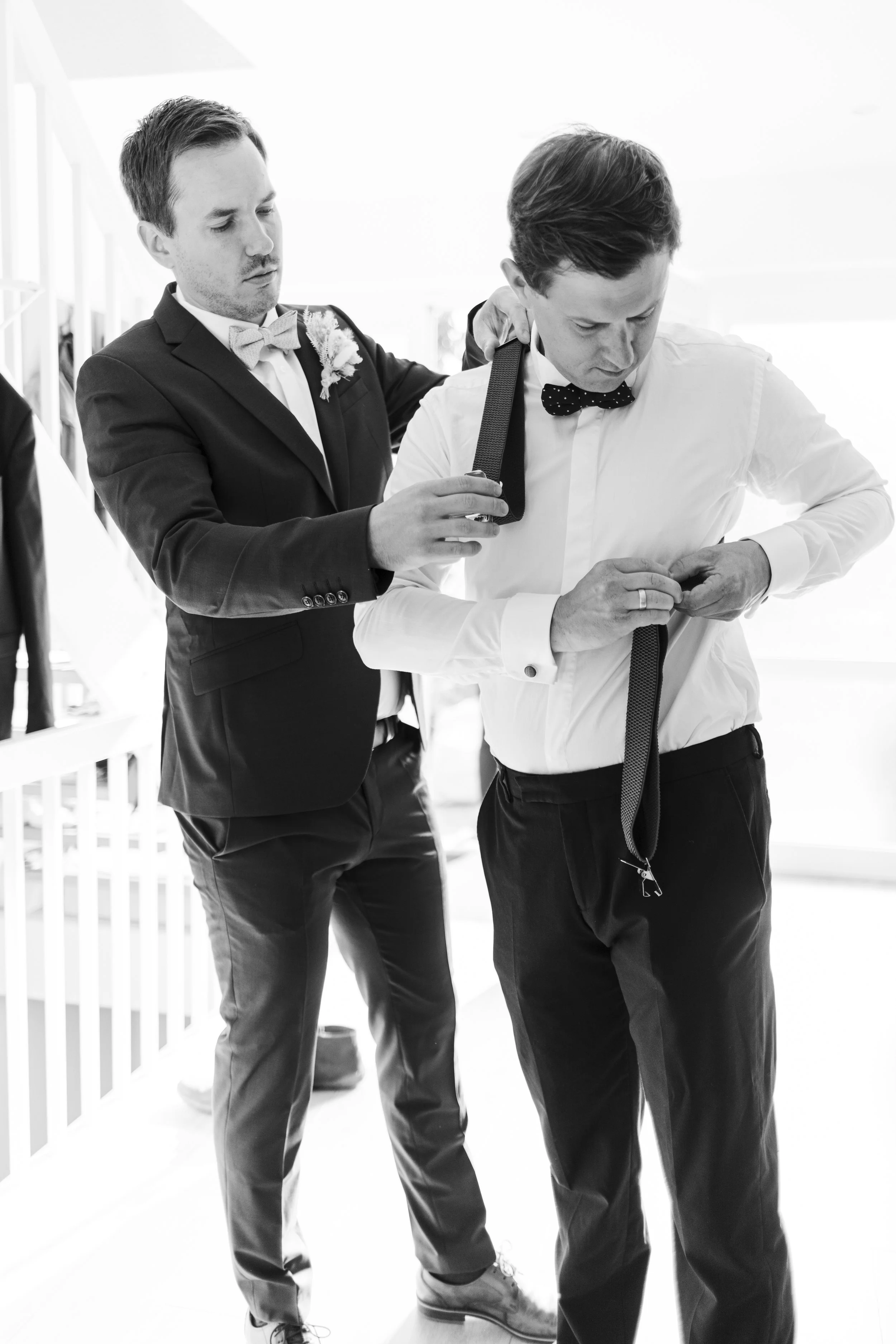 Two men dressed in formal attire, one helping the other adjust his suspenders. They are indoors with bright lighting and minimal background detail.