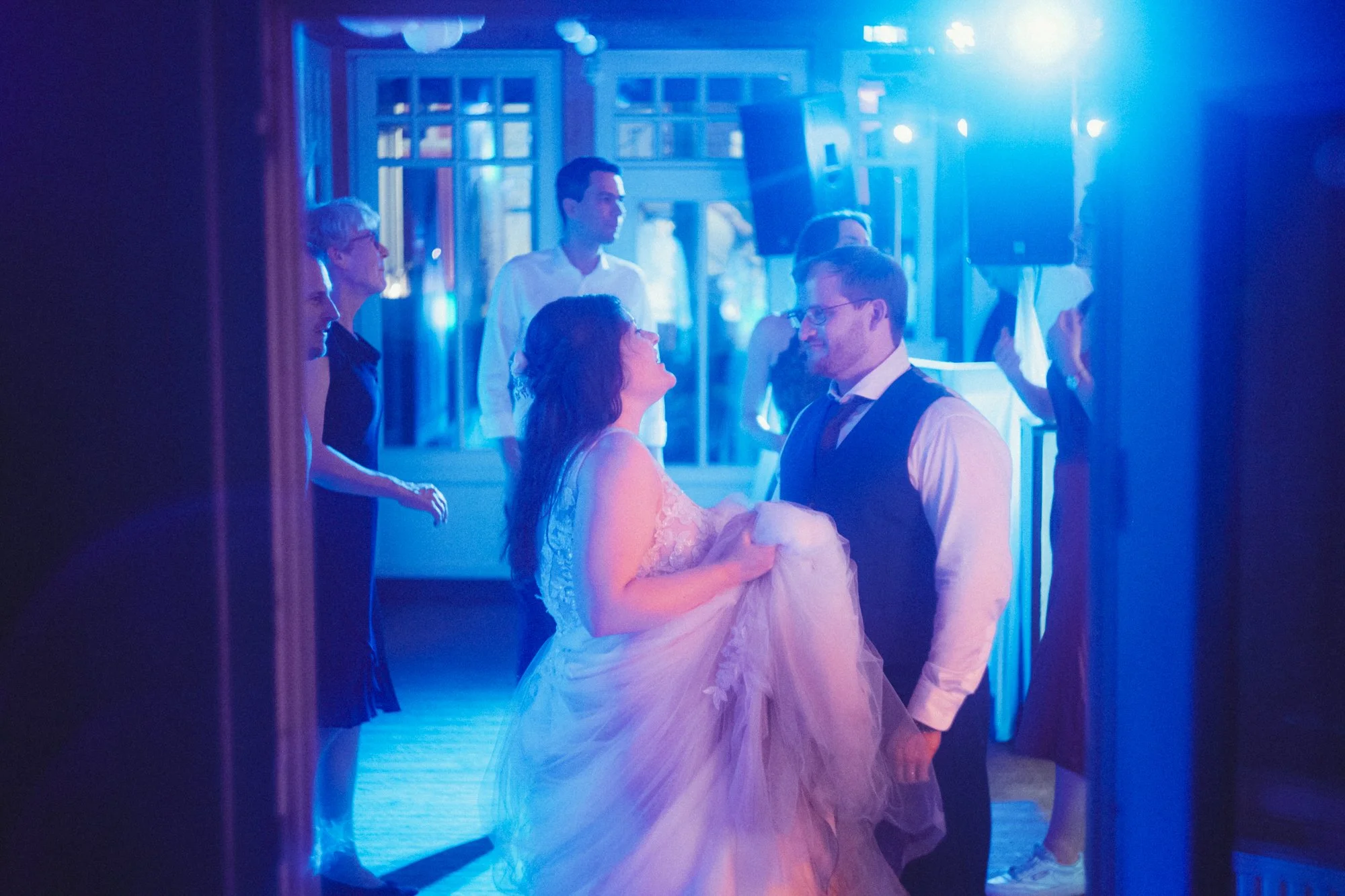 A bride and groom dance together at their wedding reception, illuminated by blue lighting, with friends and family watching nearby.