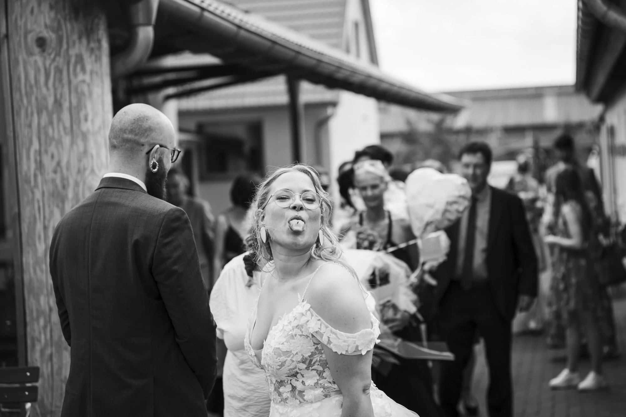 A black and white photo of a woman with glasses and a tattoo sticking out her tongue, in a wedding dress, standing next to a man in a suit, at an outdoor event with several people in the background.