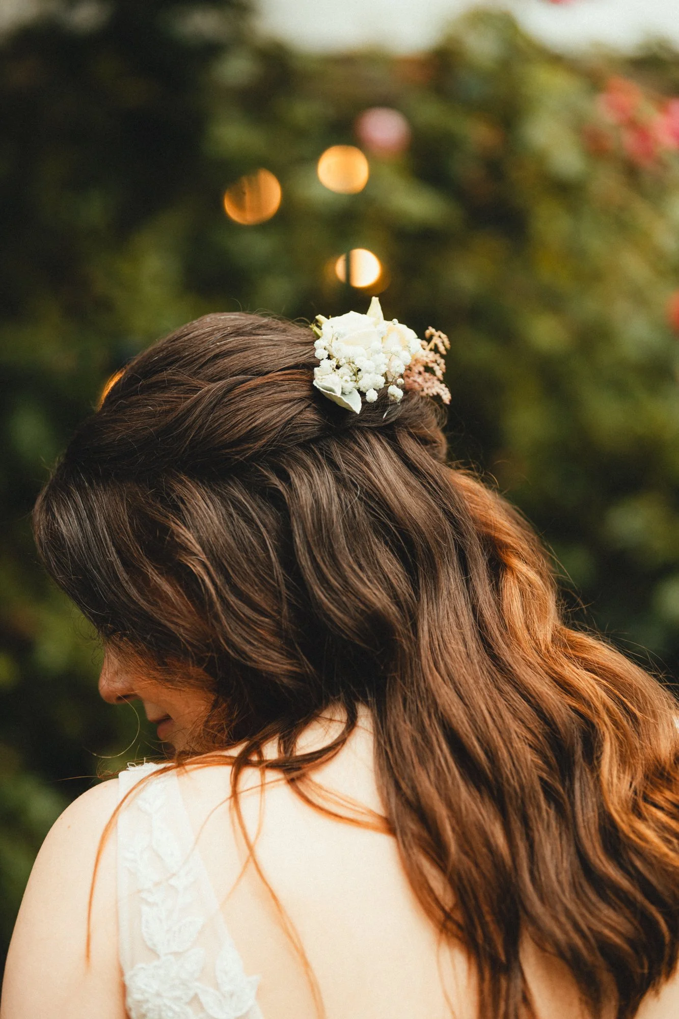 Close-up of a woman with long brown and red hair, wearing a white dress with lace details on the shoulder, and a floral hair accessory with white and light pink flowers, outdoors with blurred green foliage and warm lighting in the background.