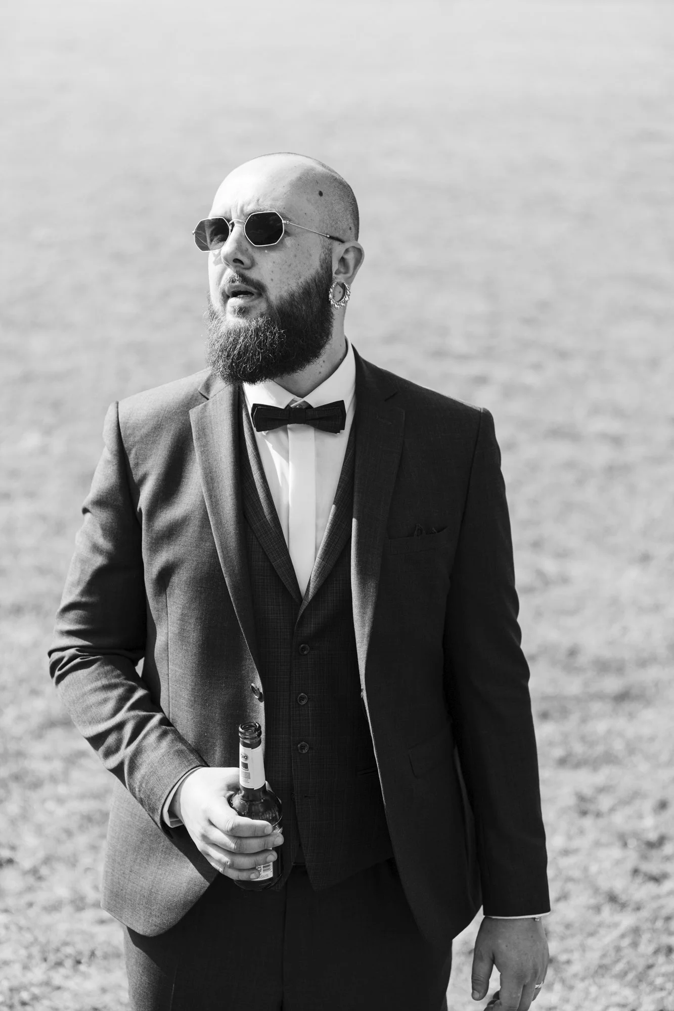 A man with a beard, a shaved head, wearing sunglasses, earrings, and a tuxedo, holding a bottle of beer while standing outdoors on a sunny day.