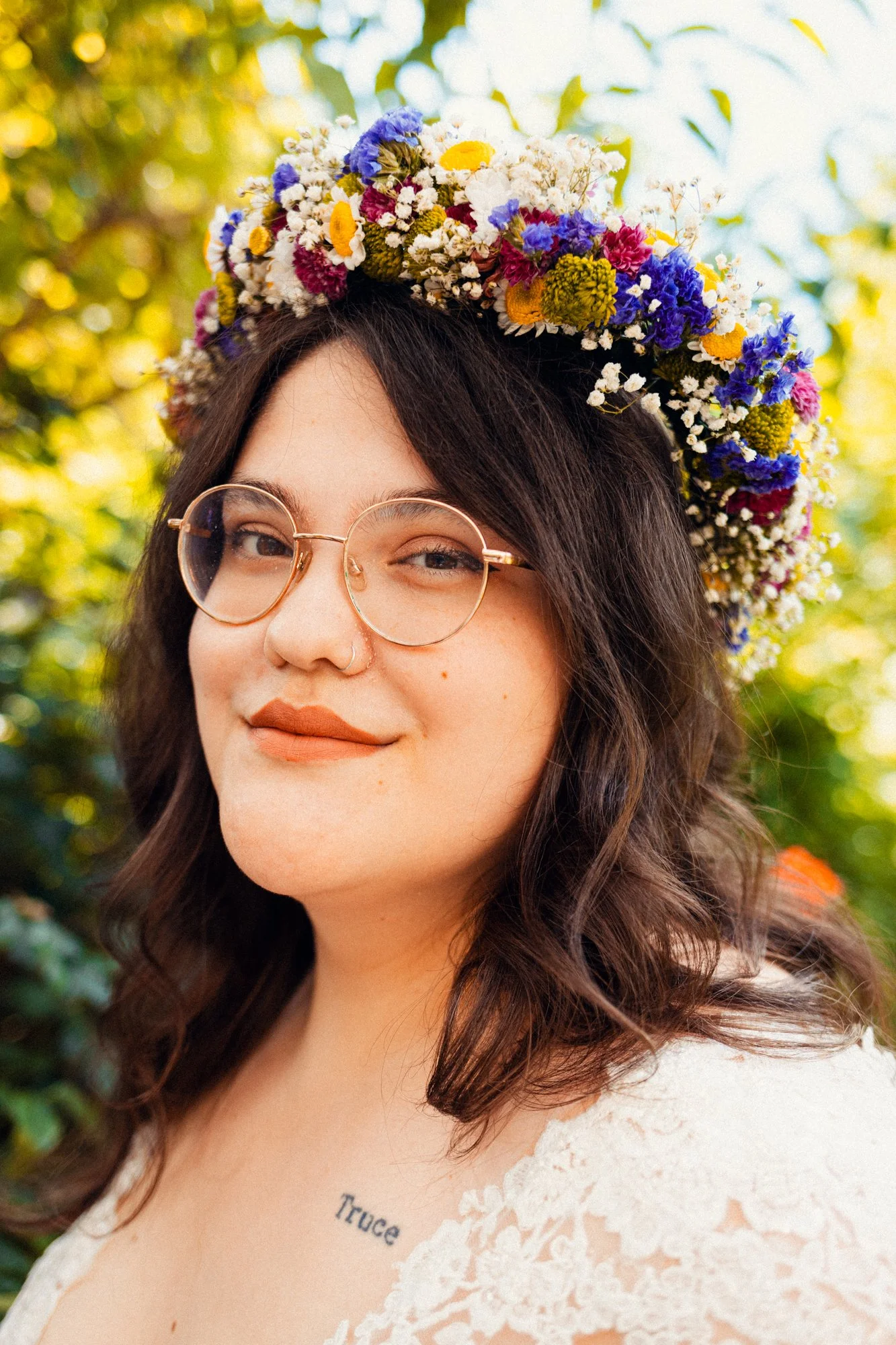A young woman with wavy brown hair, wearing glasses and a floral crown made of small, colorful flowers, outdoors with green foliage in the background.