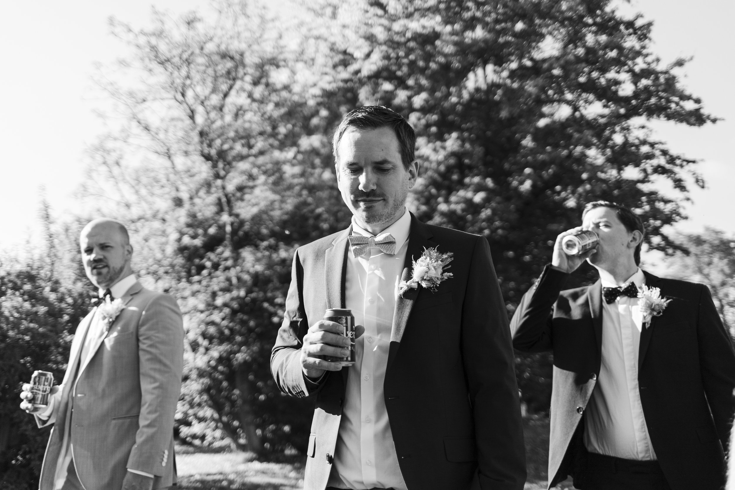 Black and white photo of three men dressed in tuxedos, holding drinks, standing outdoors with trees in the background during the daytime.