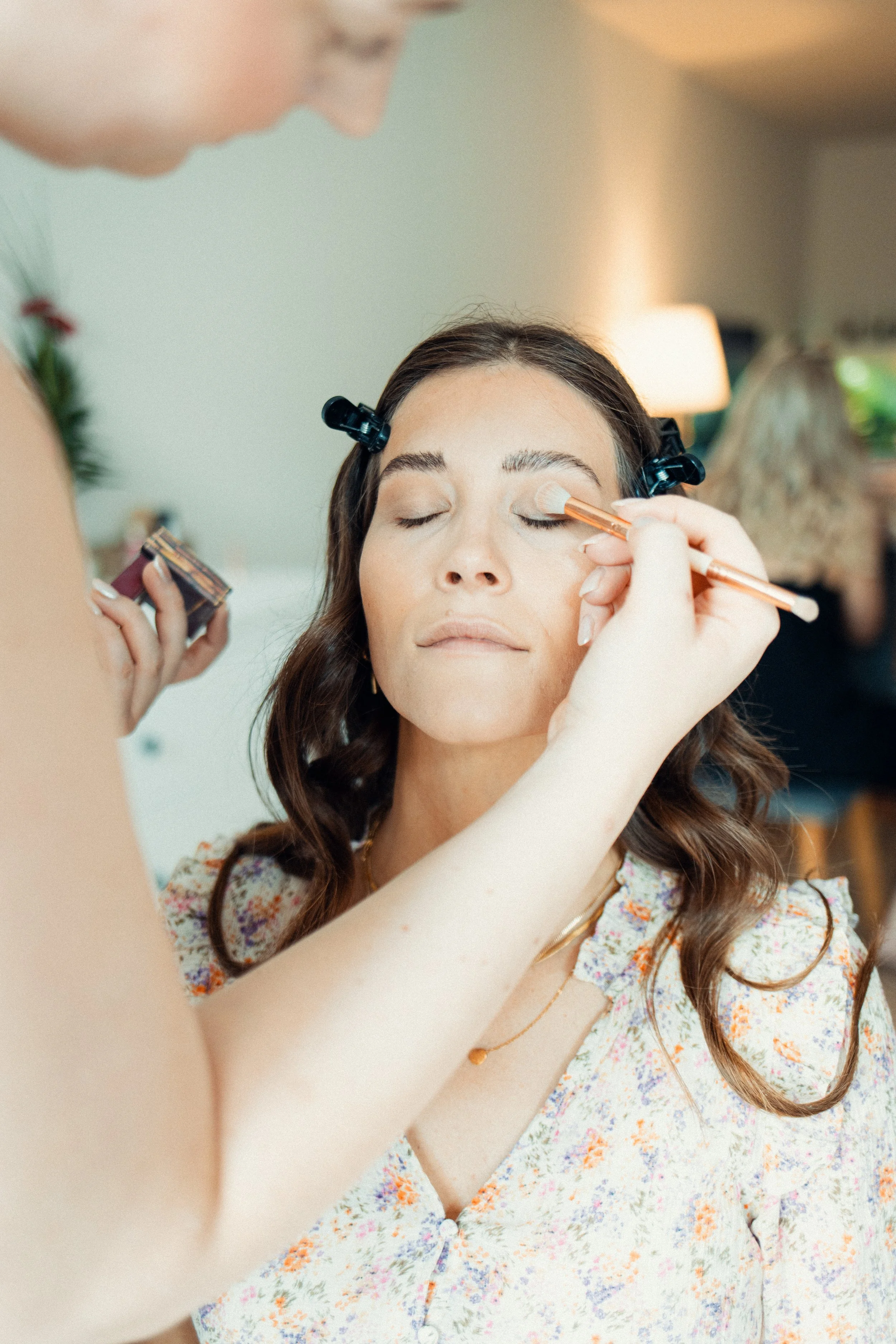 A woman with long, wavy brown hair getting her eye makeup done by a person holding a makeup brush and a palette, in a well-lit room.