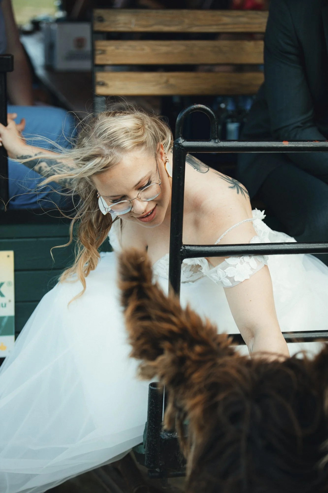 A young woman with glasses and earrings, wearing a white dress with floral details, is leaning over a white surface outdoors. Her blonde hair is blowing in the wind. Part of a dog with brown, curly fur is visible in the foreground. There are wooden b
