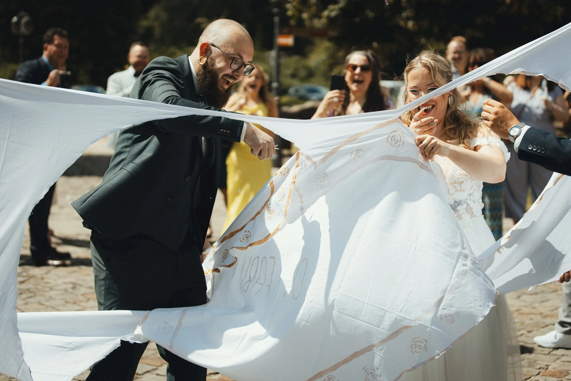 People at a wedding celebration tearing a white fabric or banner with gold roses design, smiling and laughing outside on a sunny day.
