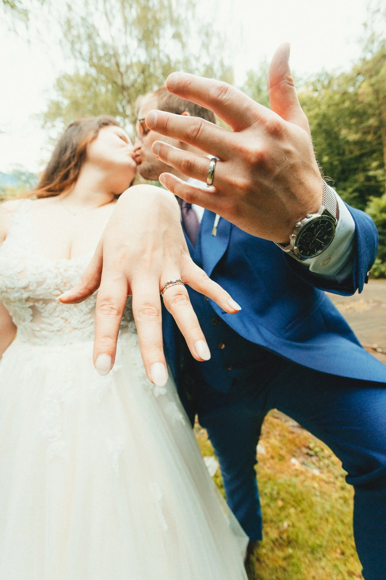A couple kissing outdoors, with their hands prominently displaying wedding rings; the woman in a wedding dress and the man in a blue suit.