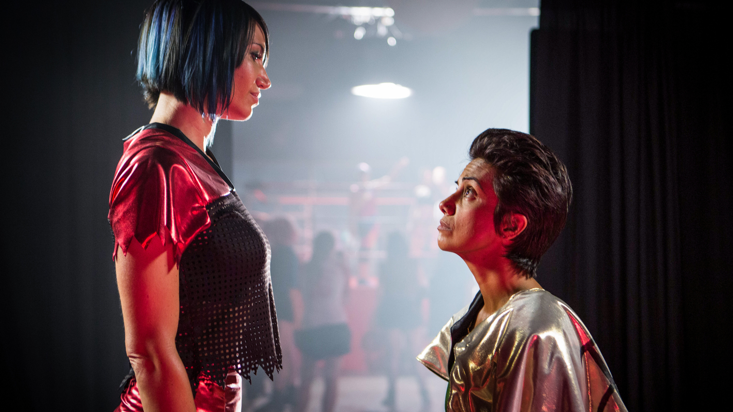 Two performers in wrestling costumes are interacting on a stage set, one standing and the other looking up while kneeling. The setting includes dark, theatrical lighting.