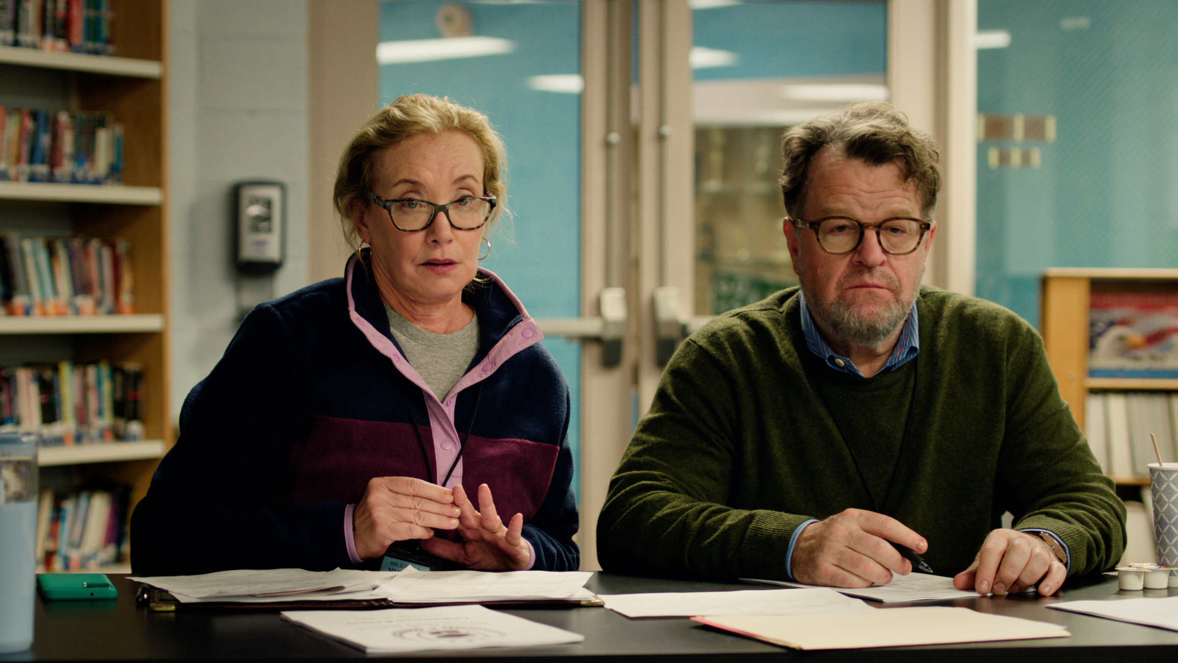 Two people sitting at a desk in a library, portraying characters from the "Only Murders in the Building" series. They are focused on paperwork in front of them.