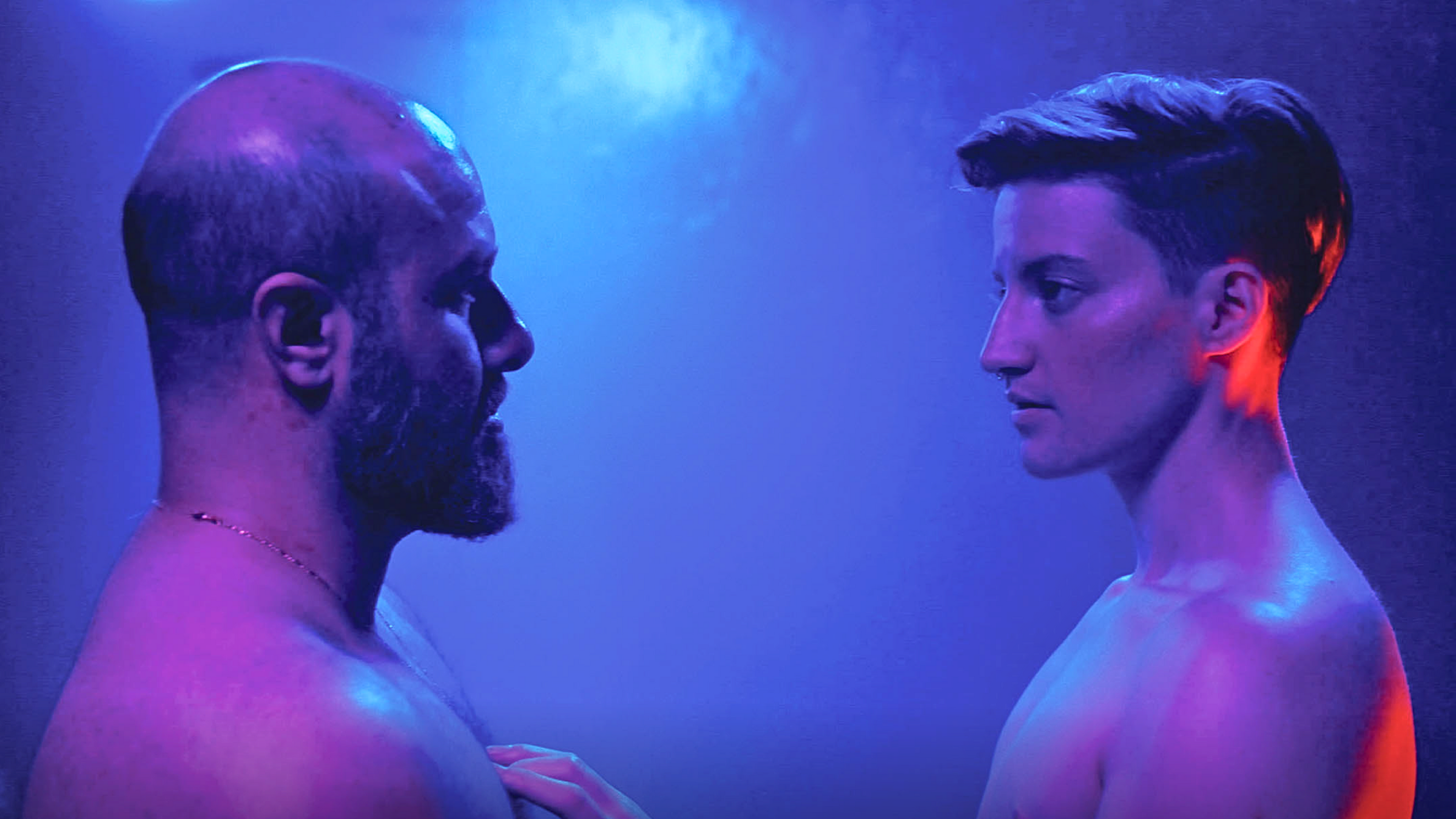 Two trans men facing each other under blue and purple lighting, conveying an intense and dramatic atmosphere.