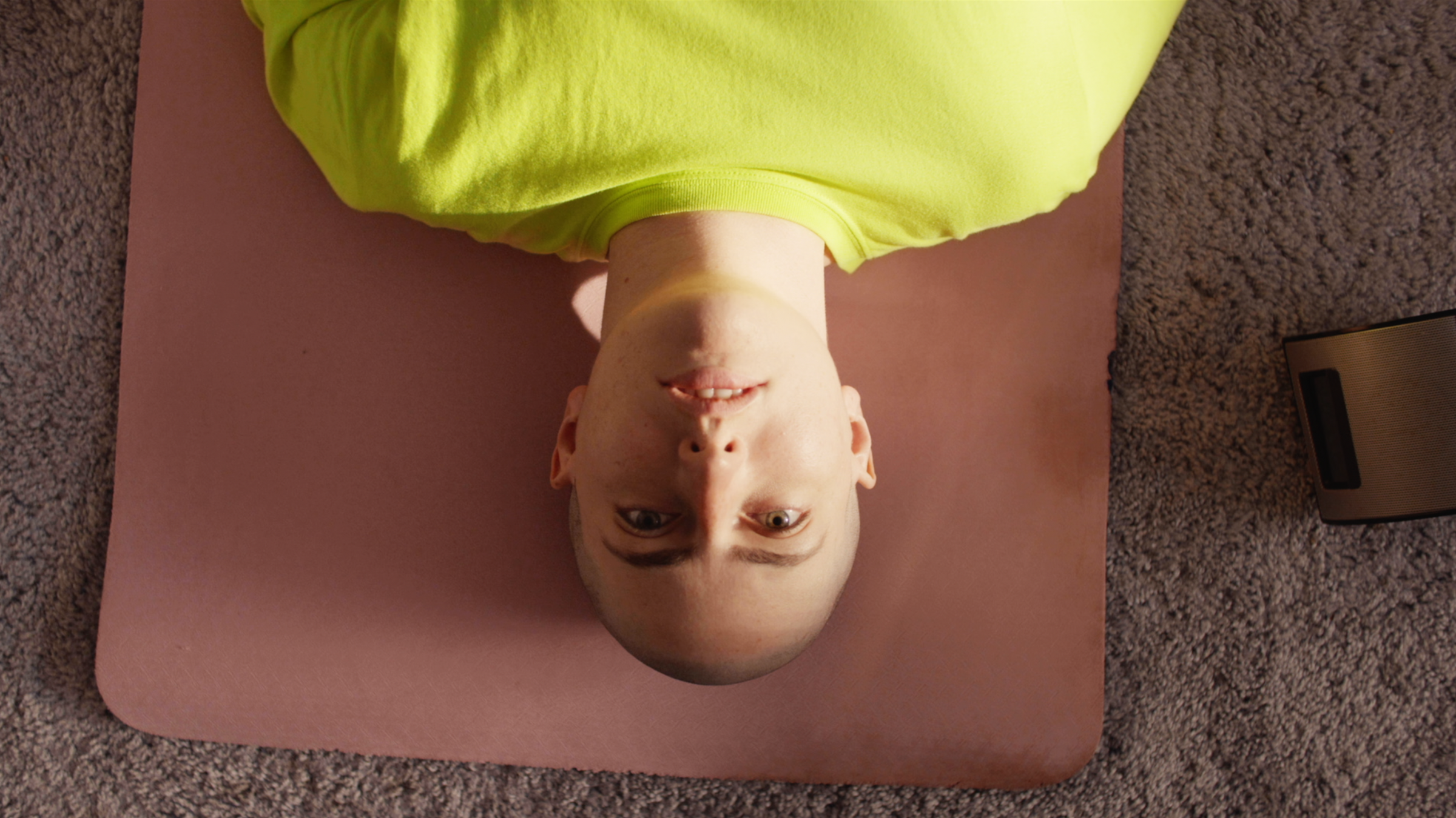 A person in a neon yellow shirt lying on a yoga mat, viewed from above, looking at the camera.