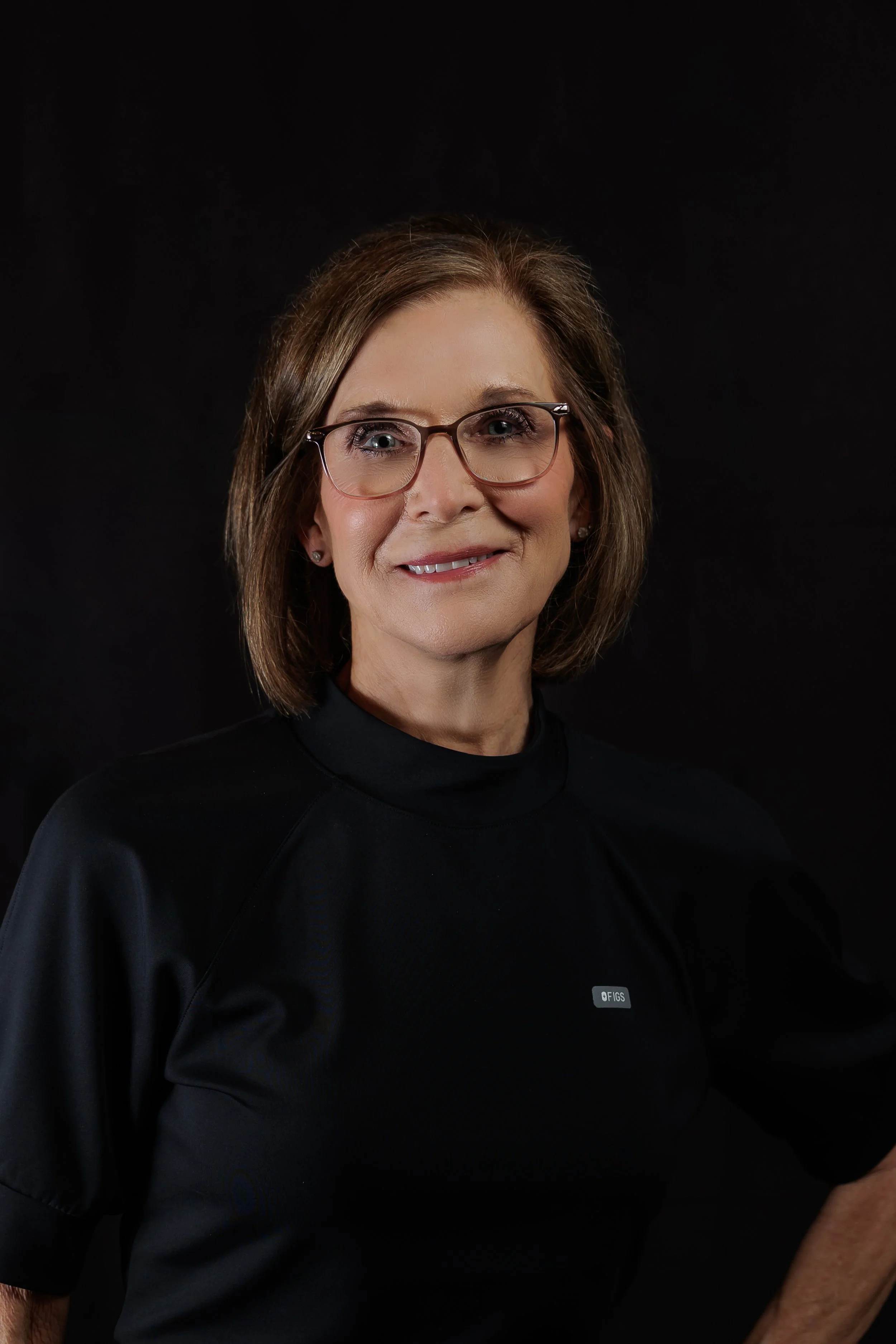 Portrait of a woman with short brown hair, glasses, and a black shirt with a small logo, smiling against a dark background.