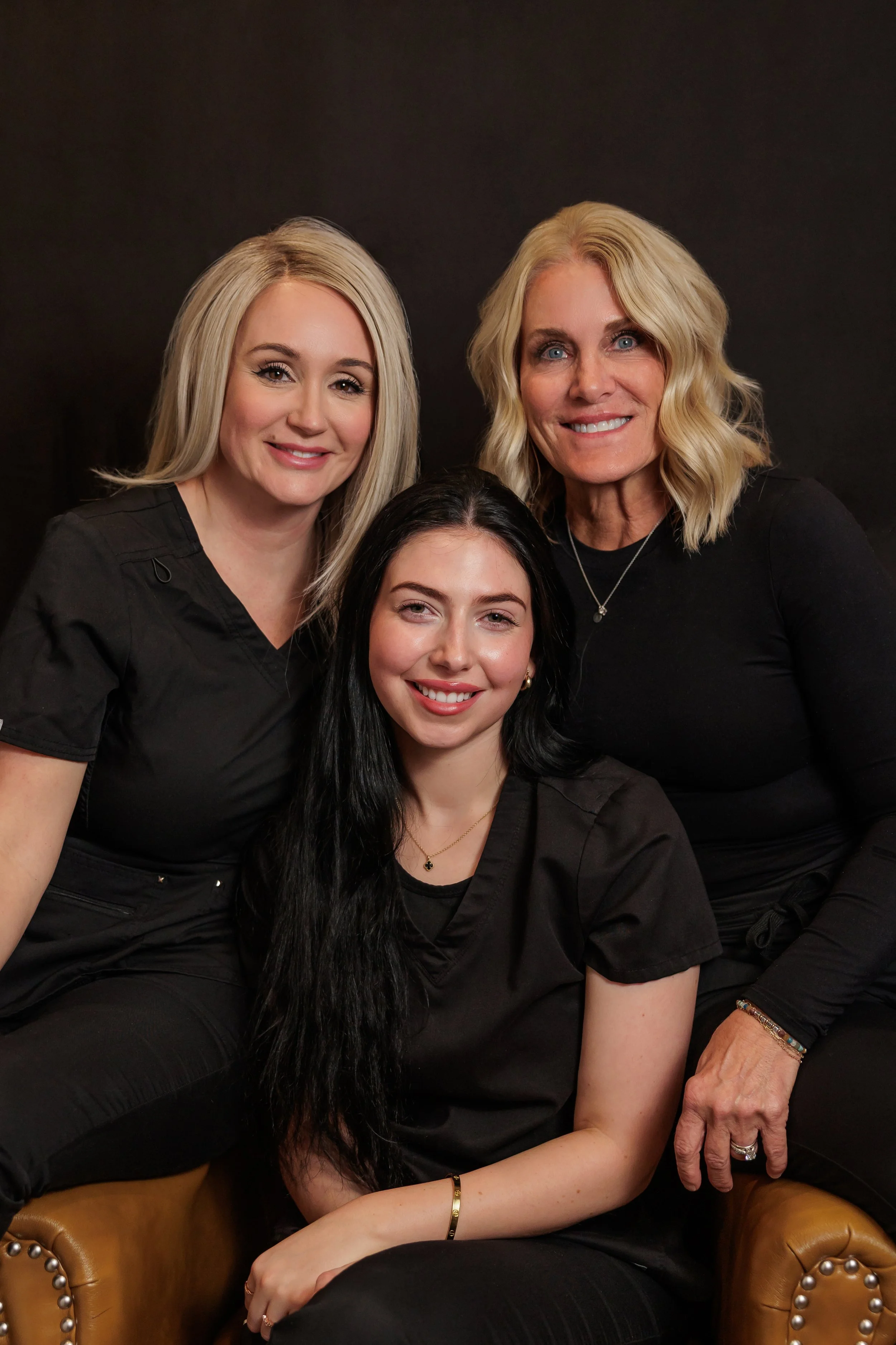Three women with blonde and black hair, smiles, wearing black clothing, posing in front of a black background.