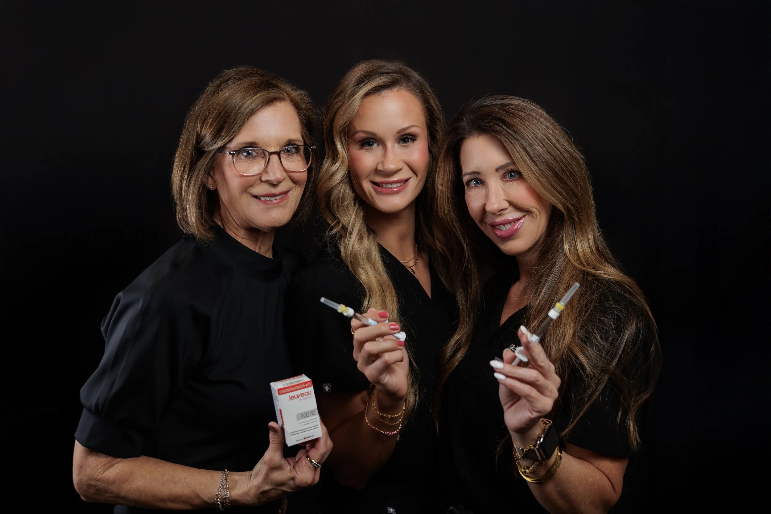 Three women smiling, holding medical syringes and medication, against a black background.